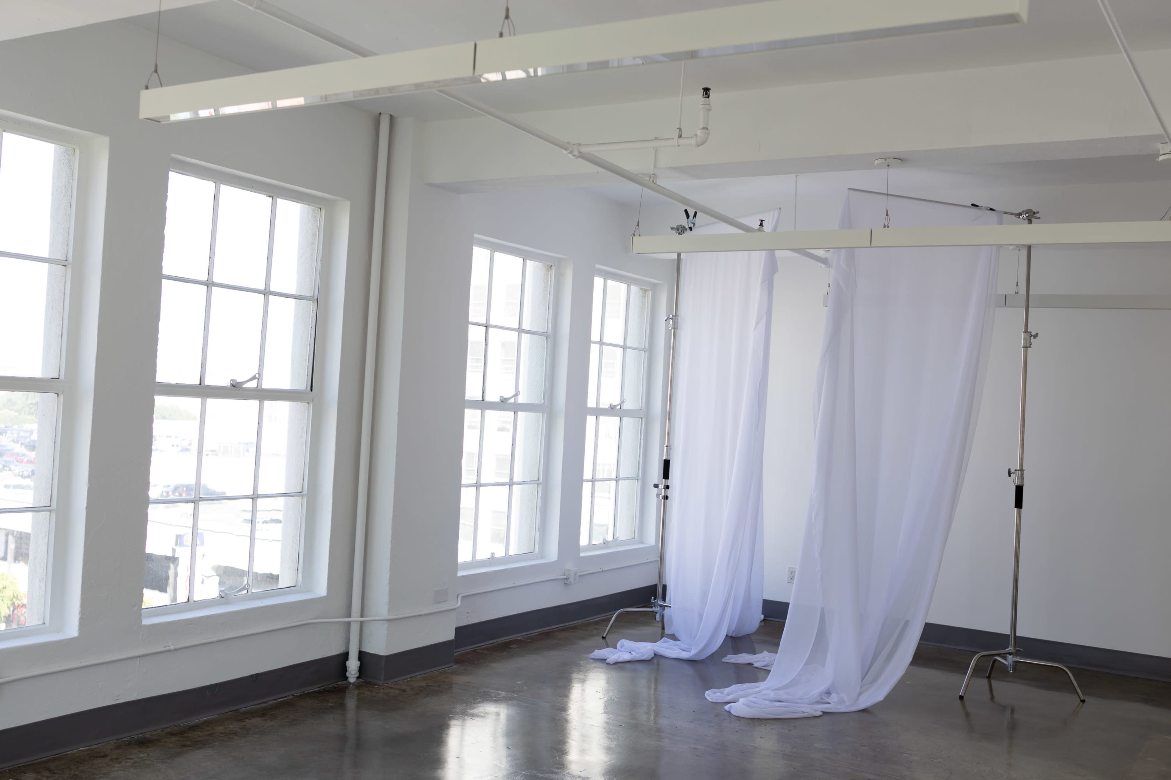 The image shows a bright, minimalist room with large windows and a sheer white curtain suspended on metal stands.