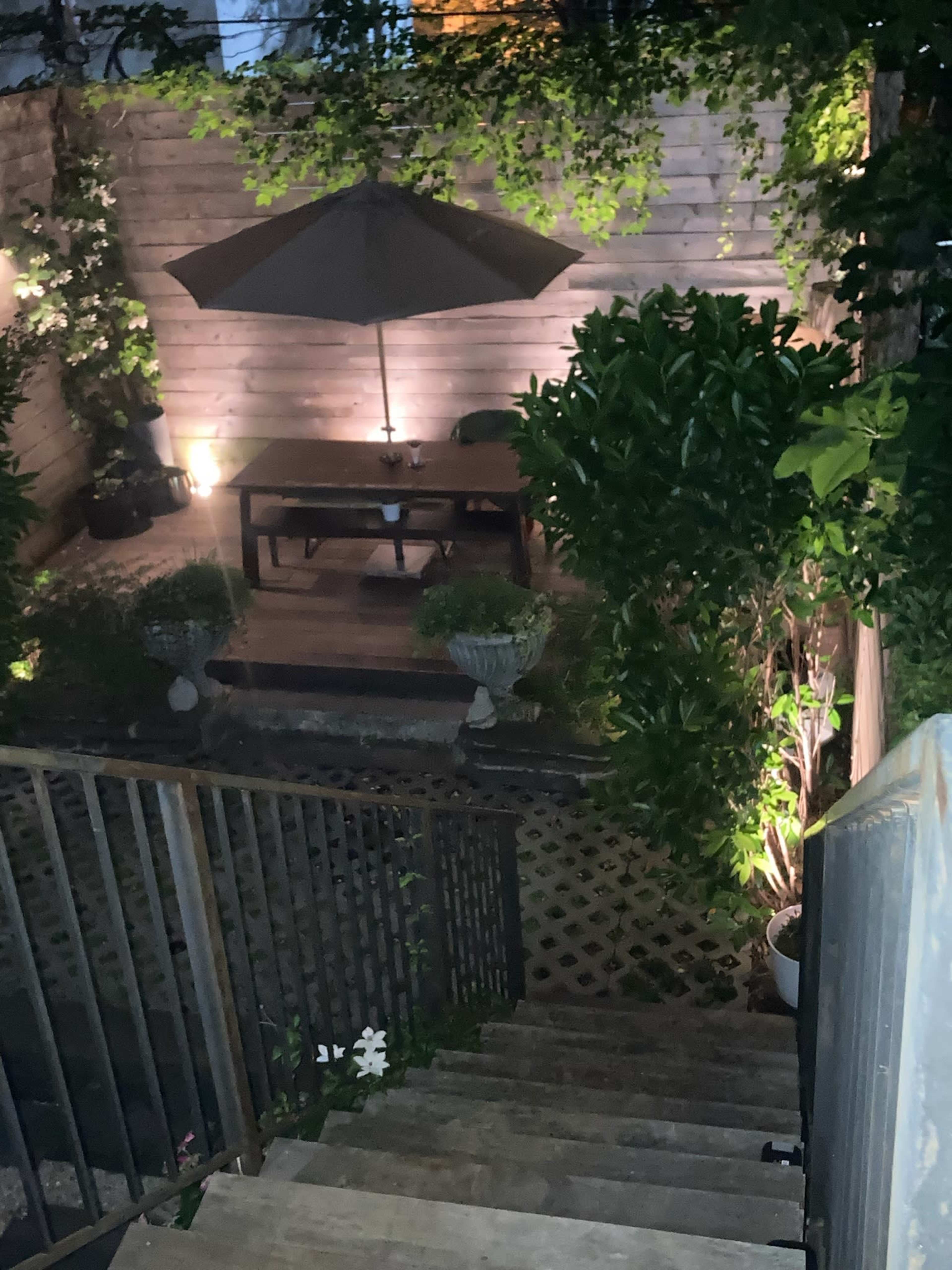 A wooden staircase leads down to a dimly lit backyard with a table and umbrella surrounded by plants.