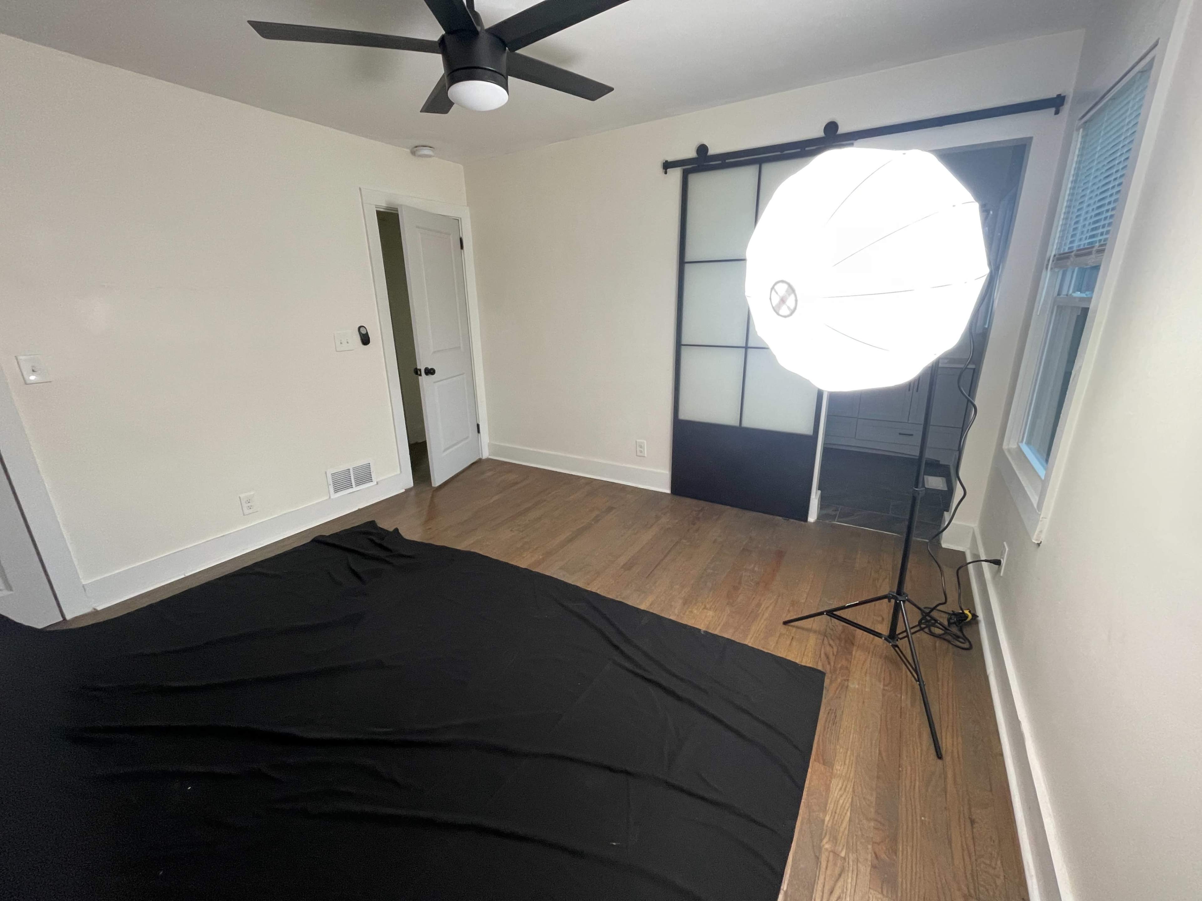 The image shows a sparsely furnished room with a black fabric on the floor and a large softbox light set up in one corner.