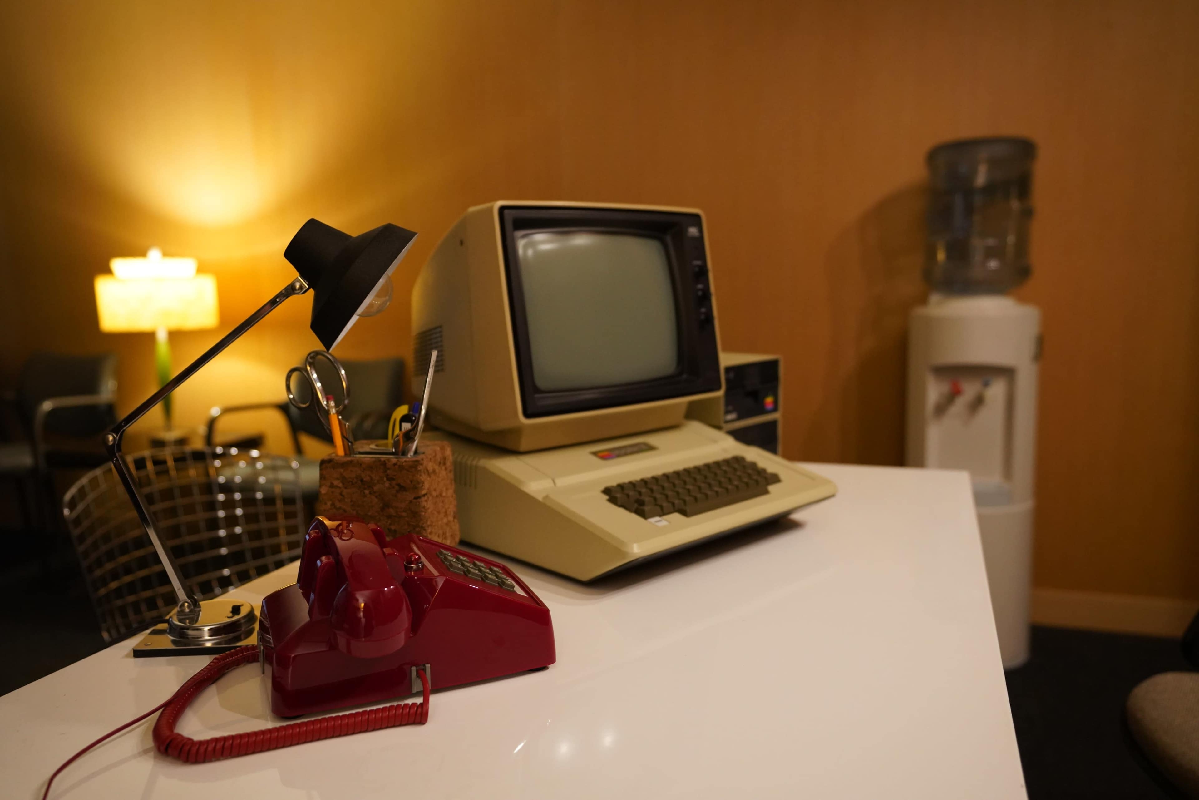 The image features a vintage computer and a red rotary telephone on a modern white desk, accompanied by a desk lamp and a water cooler in a softly lit office space.