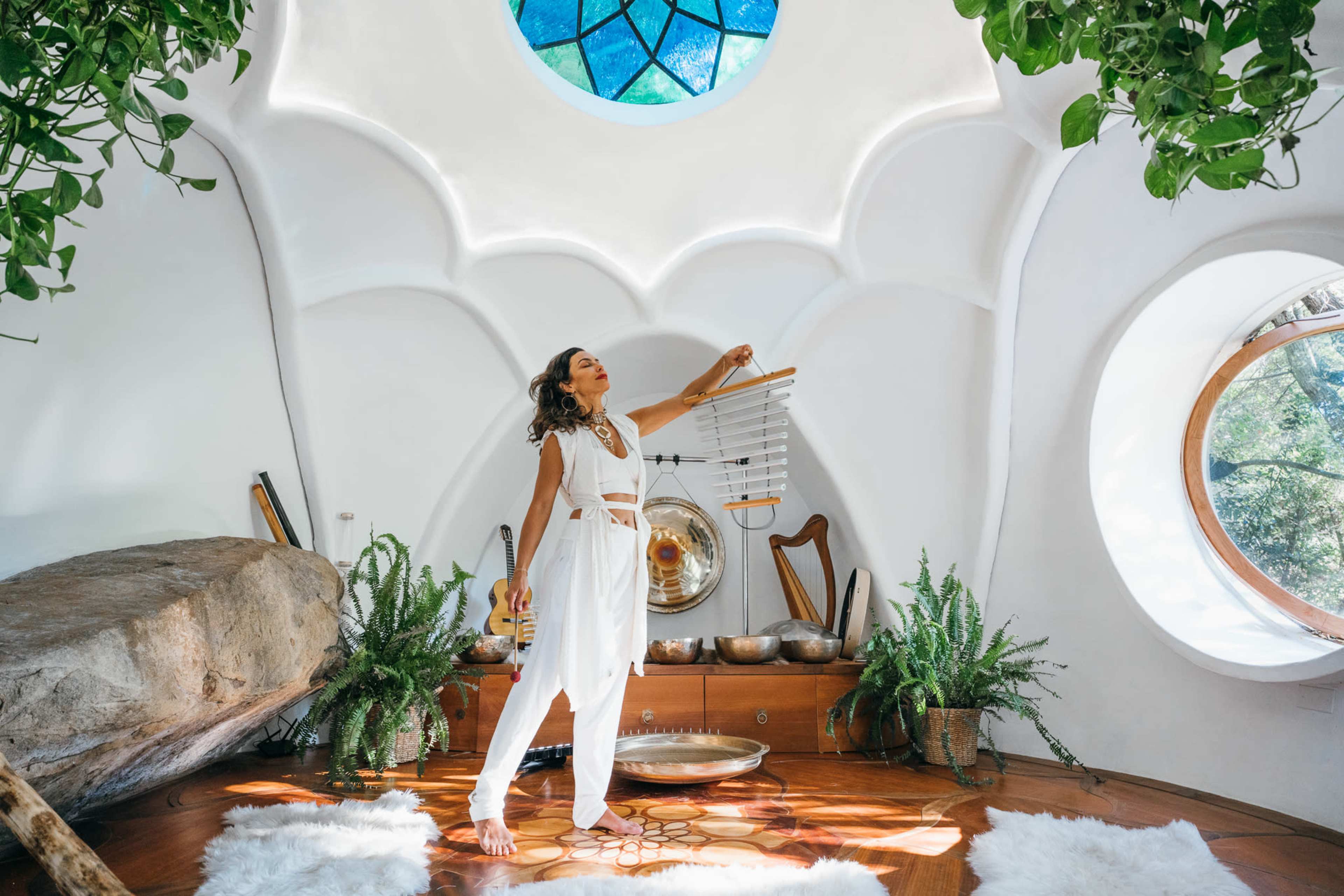 A woman stands in a brightly lit, uniquely shaped room with white walls and a large circular window, holding a musical chime while surrounded by plants and decorative objects.