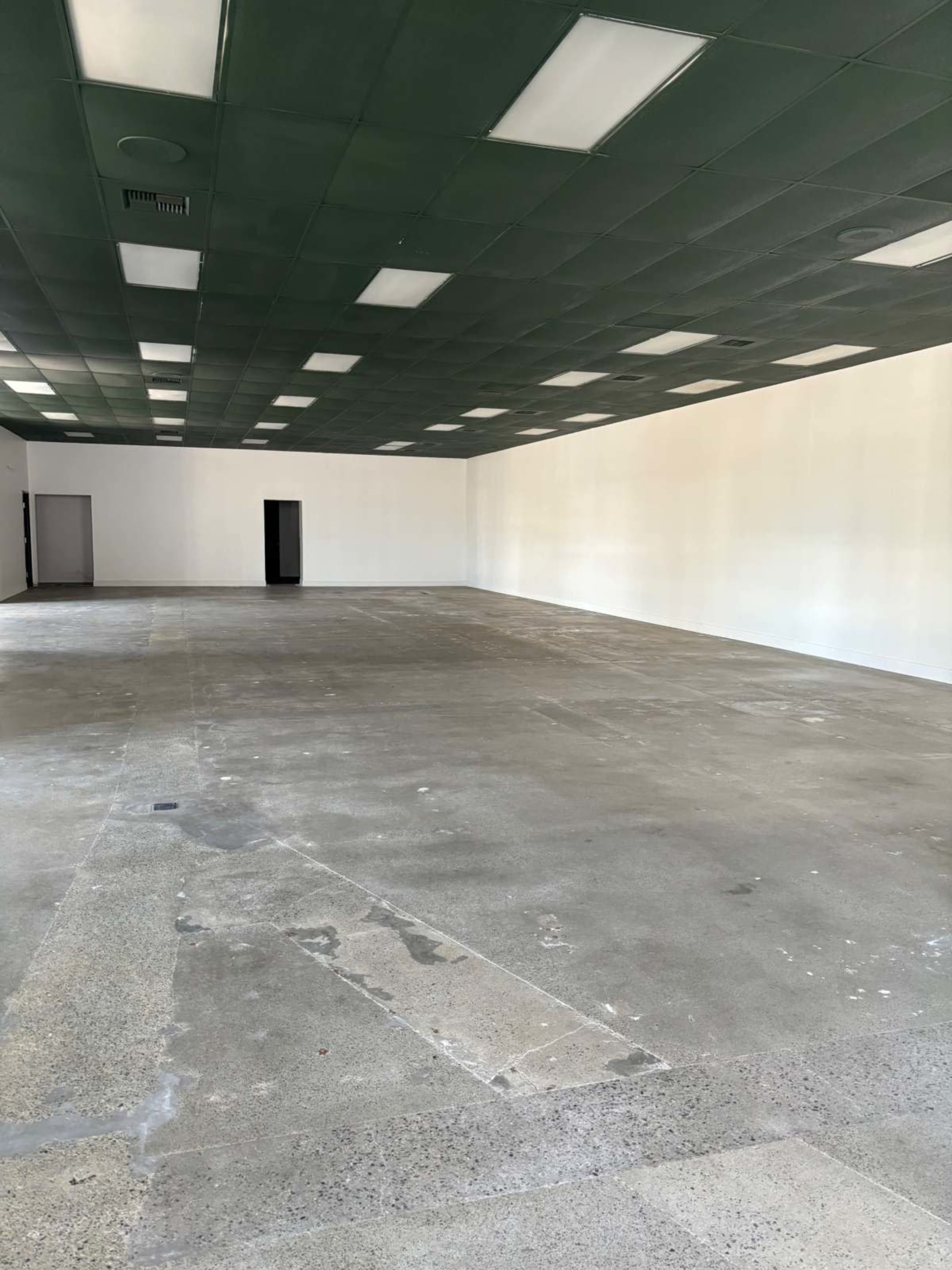 The image shows a large, empty commercial space with a concrete floor, green ceiling tiles, and white walls, lacking any furniture or decoration.