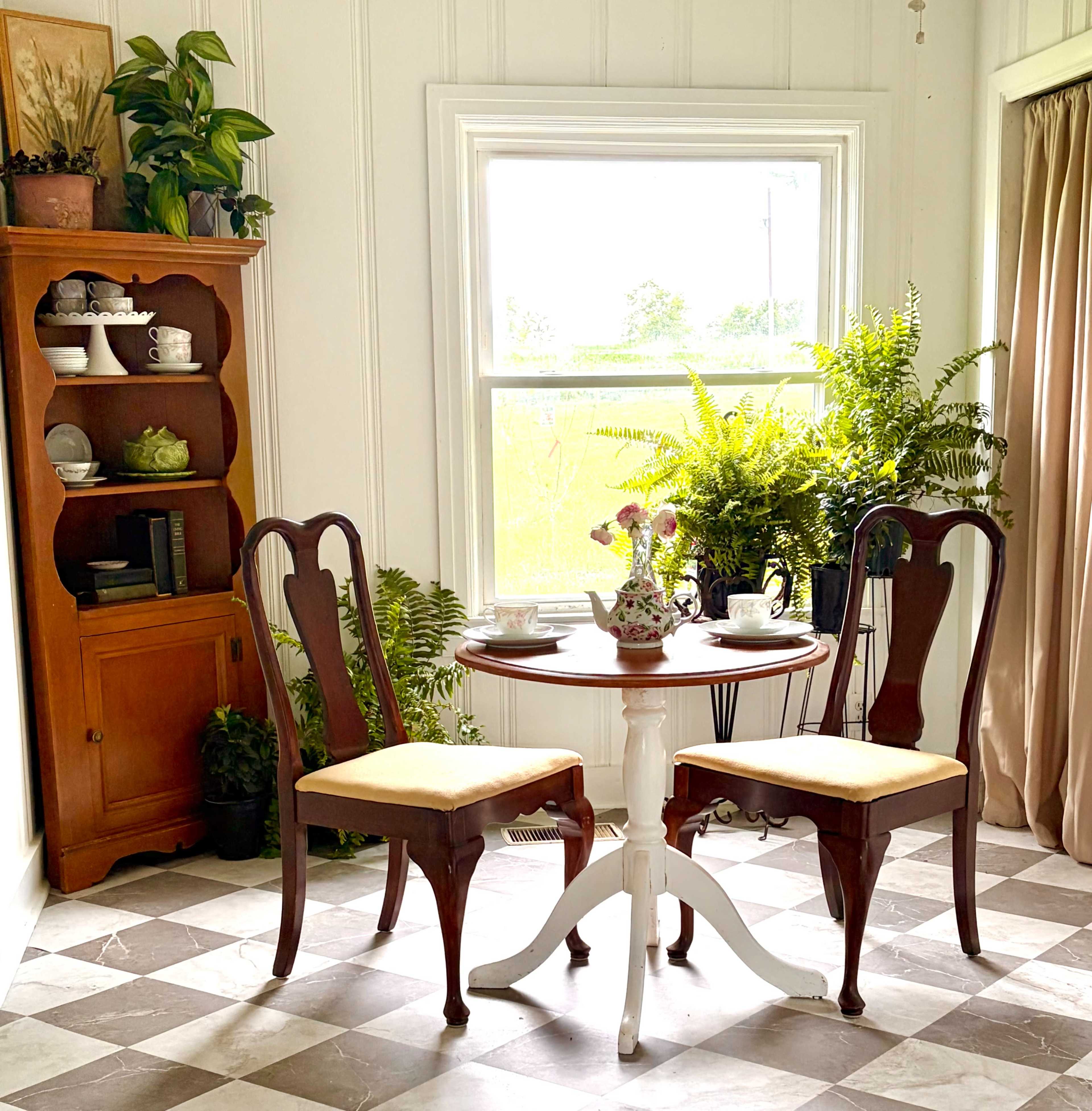 A small dining table with two chairs is positioned near a window, surrounded by plants and a wooden cabinet.