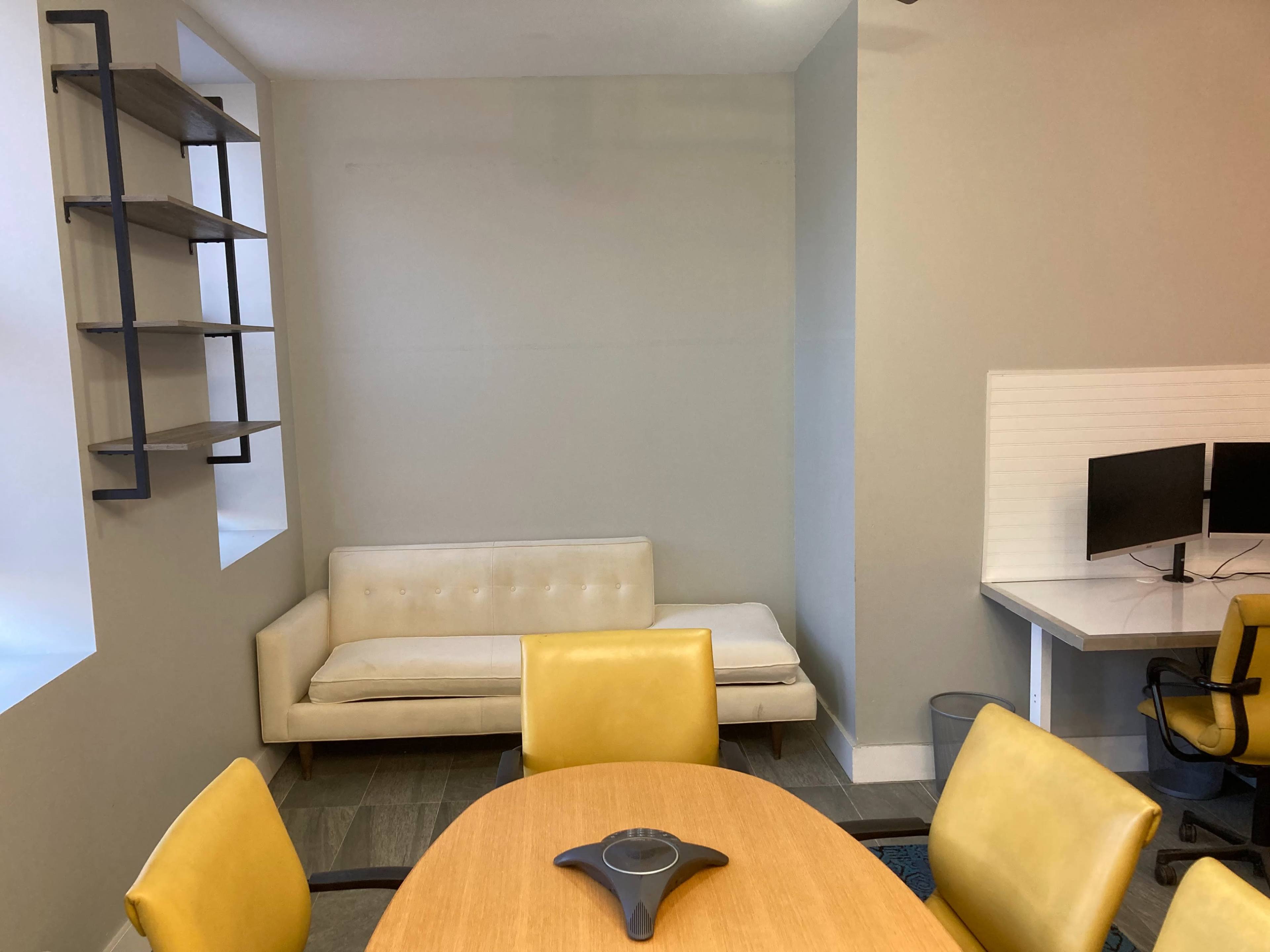 A minimalist office space with a beige couch, a round table, yellow chairs, and two computer monitors on a desk.