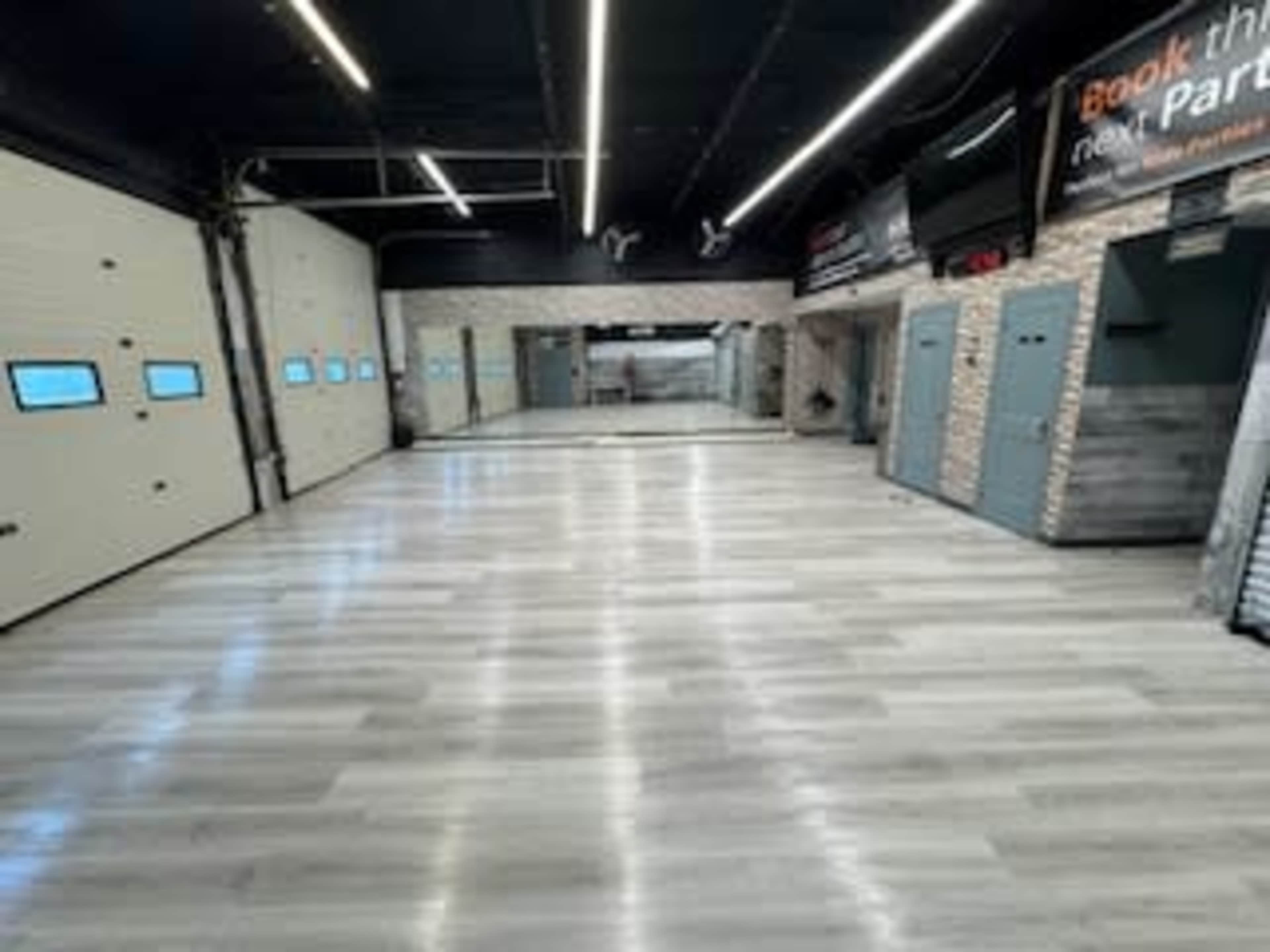The image shows a spacious indoor area with a smooth, light-colored wooden floor, illuminated by overhead lights, featuring large garage-style doors and mirrored walls.