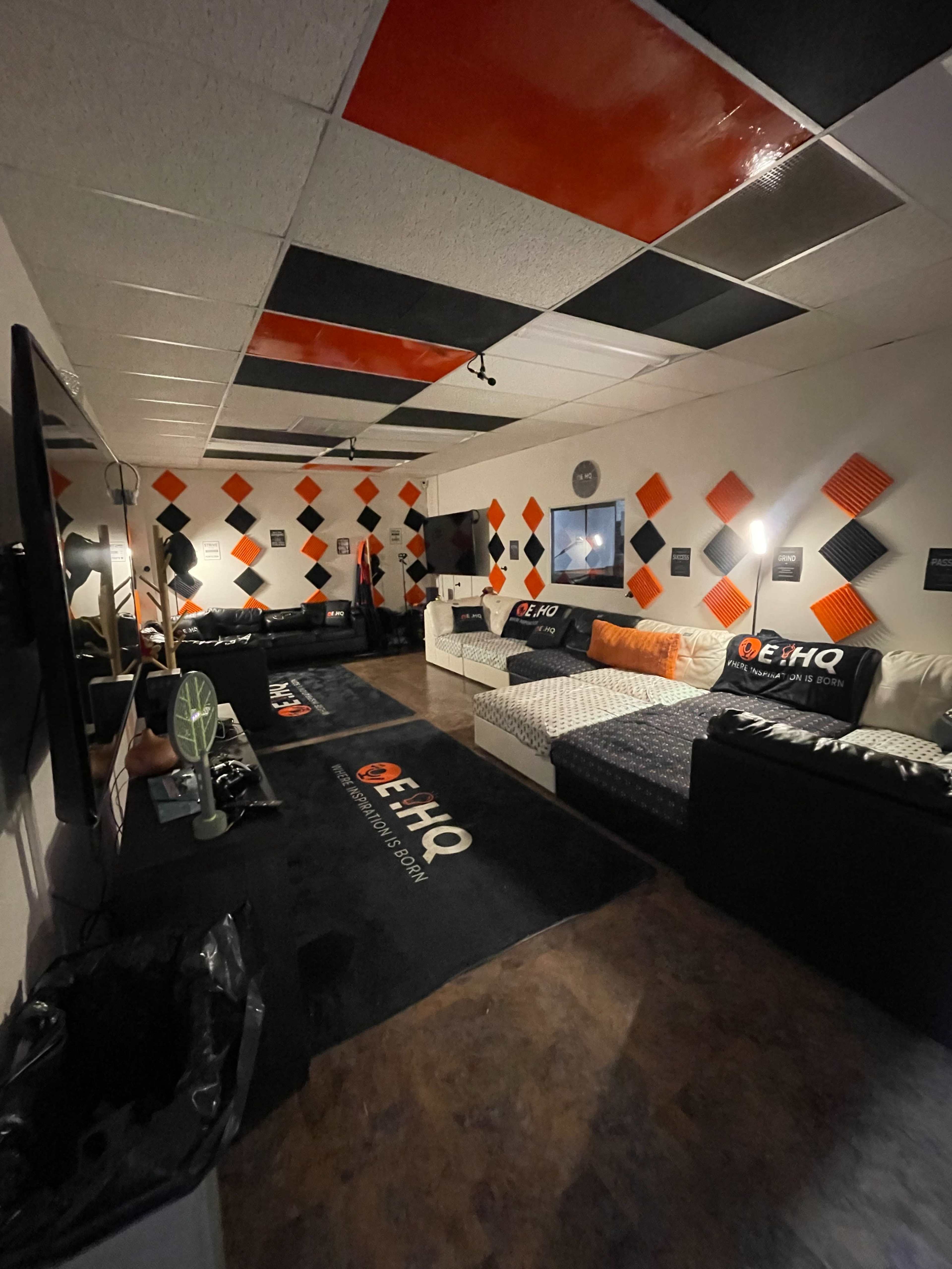 The image shows a room with black and orange decor, featuring couches, a television, and patterned acoustic panels on the walls.