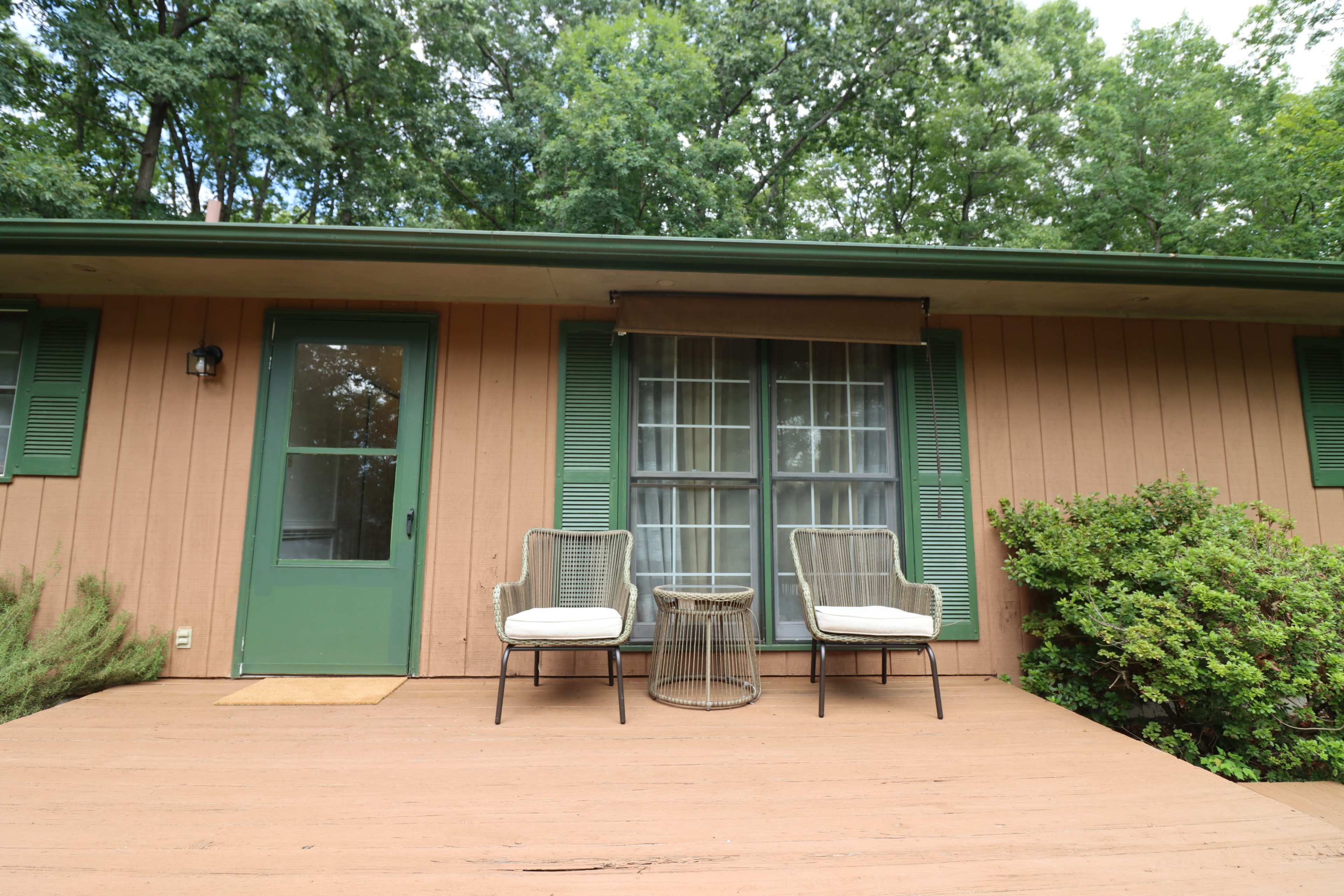 A wooden porch with two chairs and a small table in front of a house featuring green shutters and a door.