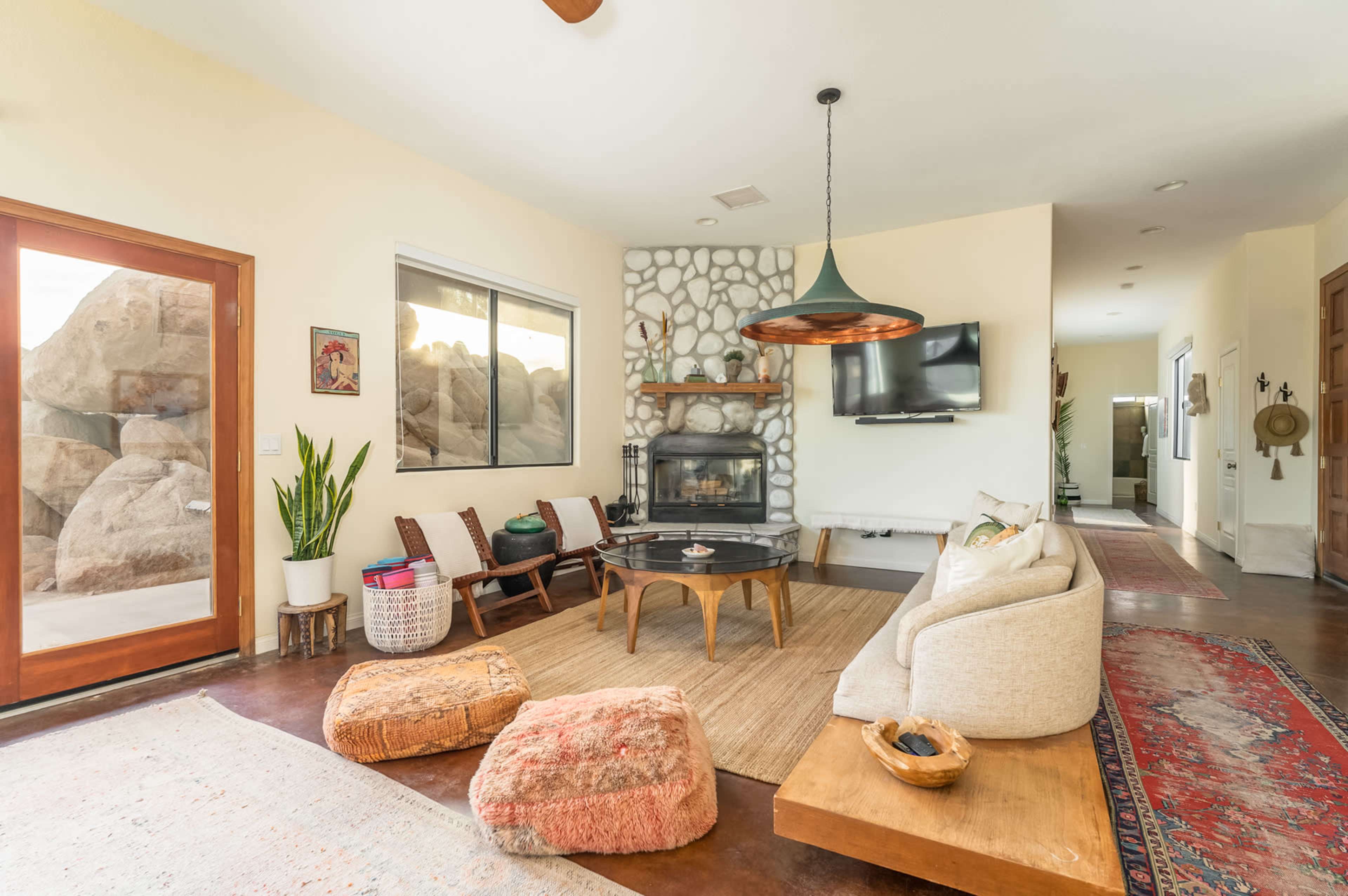 The interior of a rustic living room features a stone fireplace, a comfortable sofa, a round wooden coffee table, and large windows showcasing boulders outside.