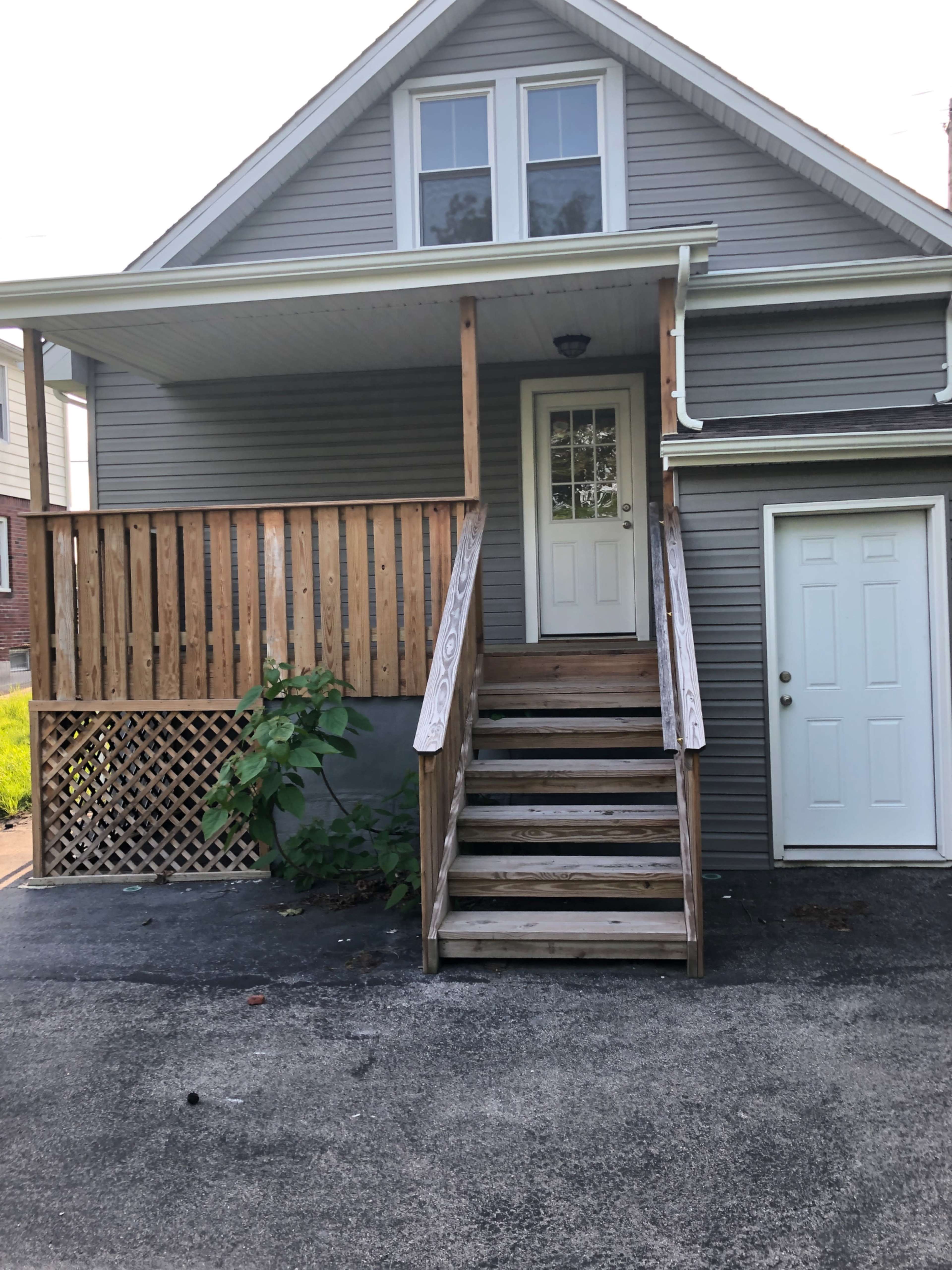 The image shows a gray house with a front porch, wooden steps leading to a white door, and a small deck area with a lattice design.