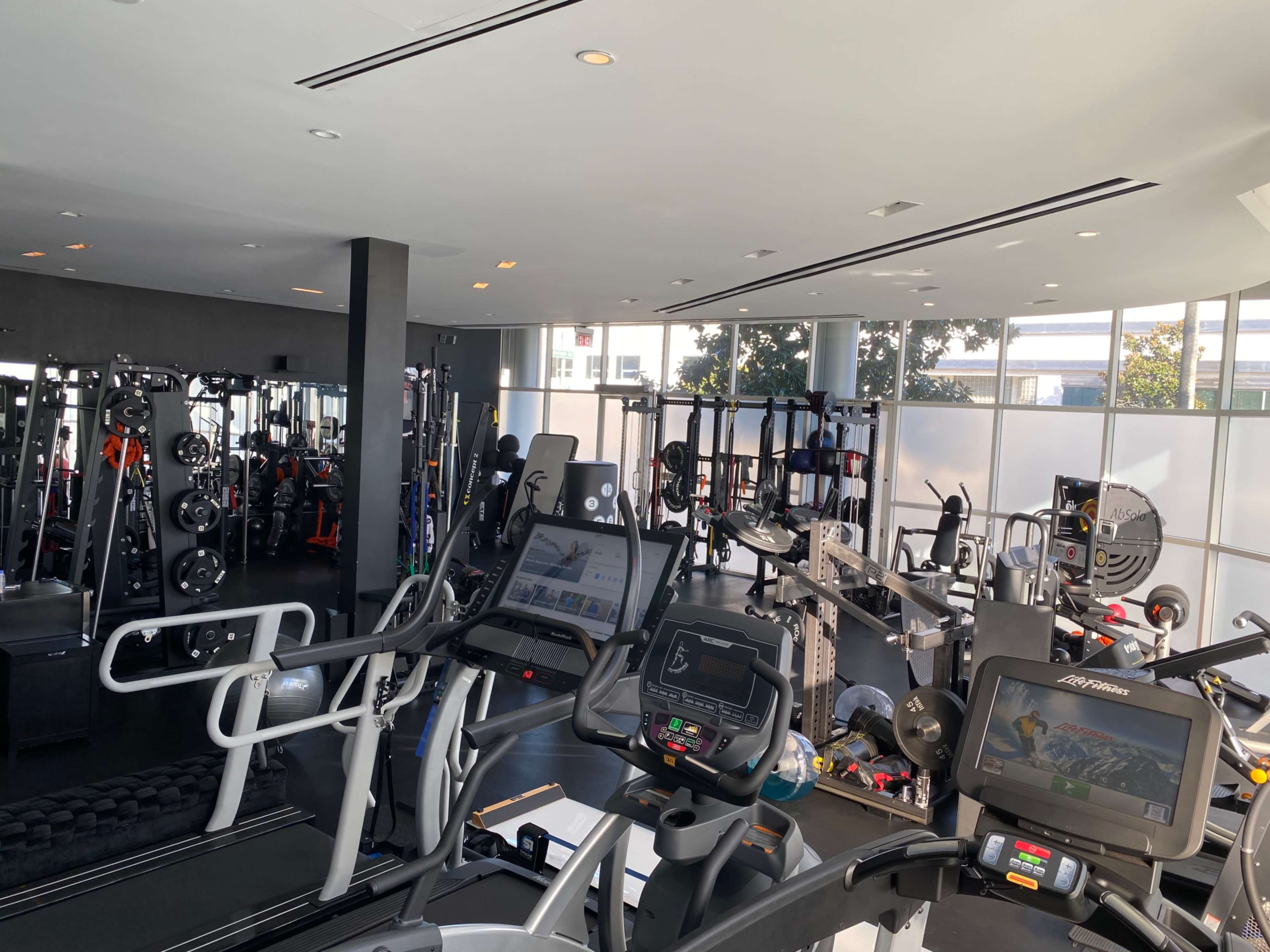 The image shows a modern gym filled with various exercise equipment, including treadmills, weight machines, and free weights, all in a well-lit space with large windows.
