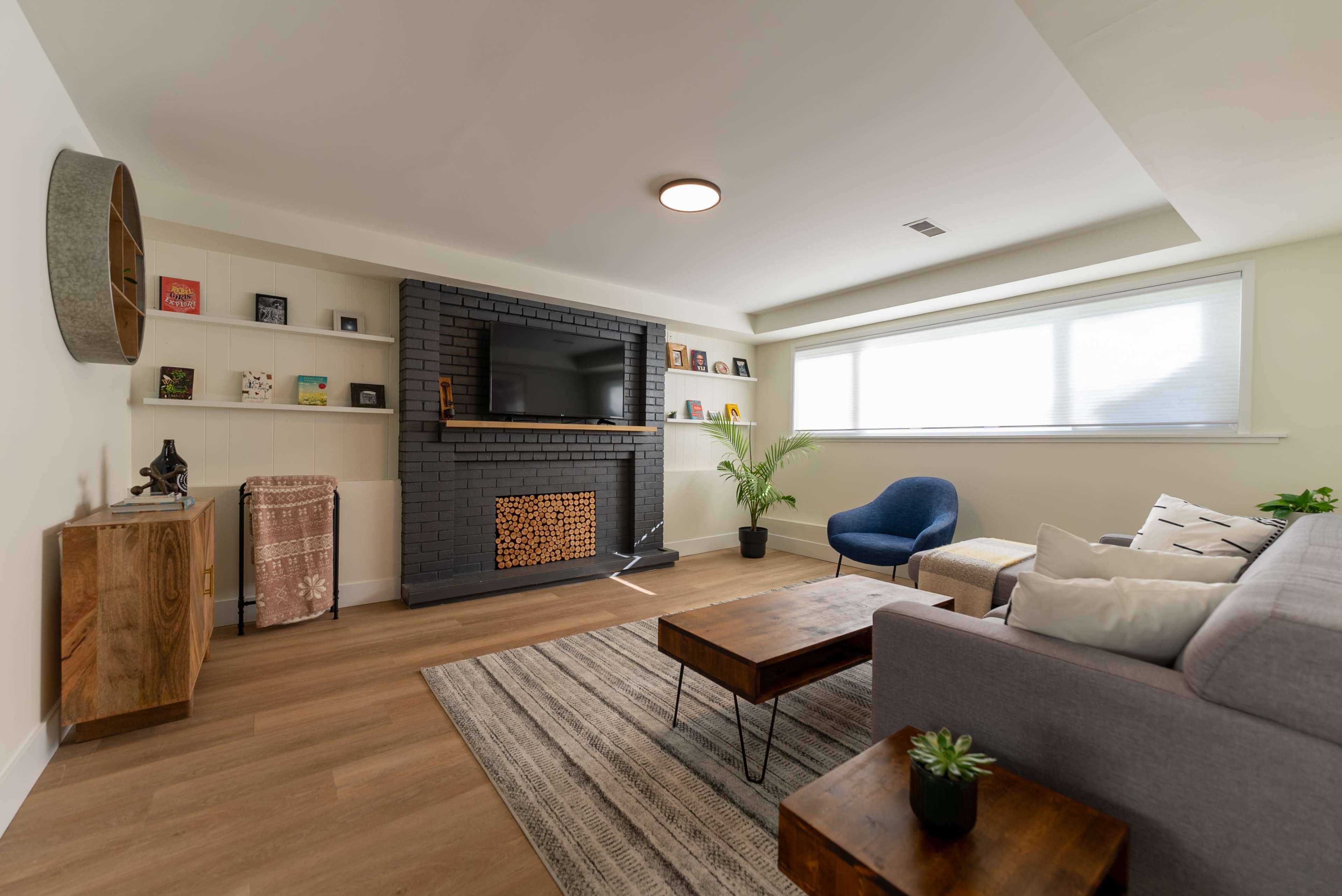 A modern living room with a gray sectional sofa, a wooden coffee table, a flat-screen TV mounted on a black brick wall, and a large window that provides natural light.