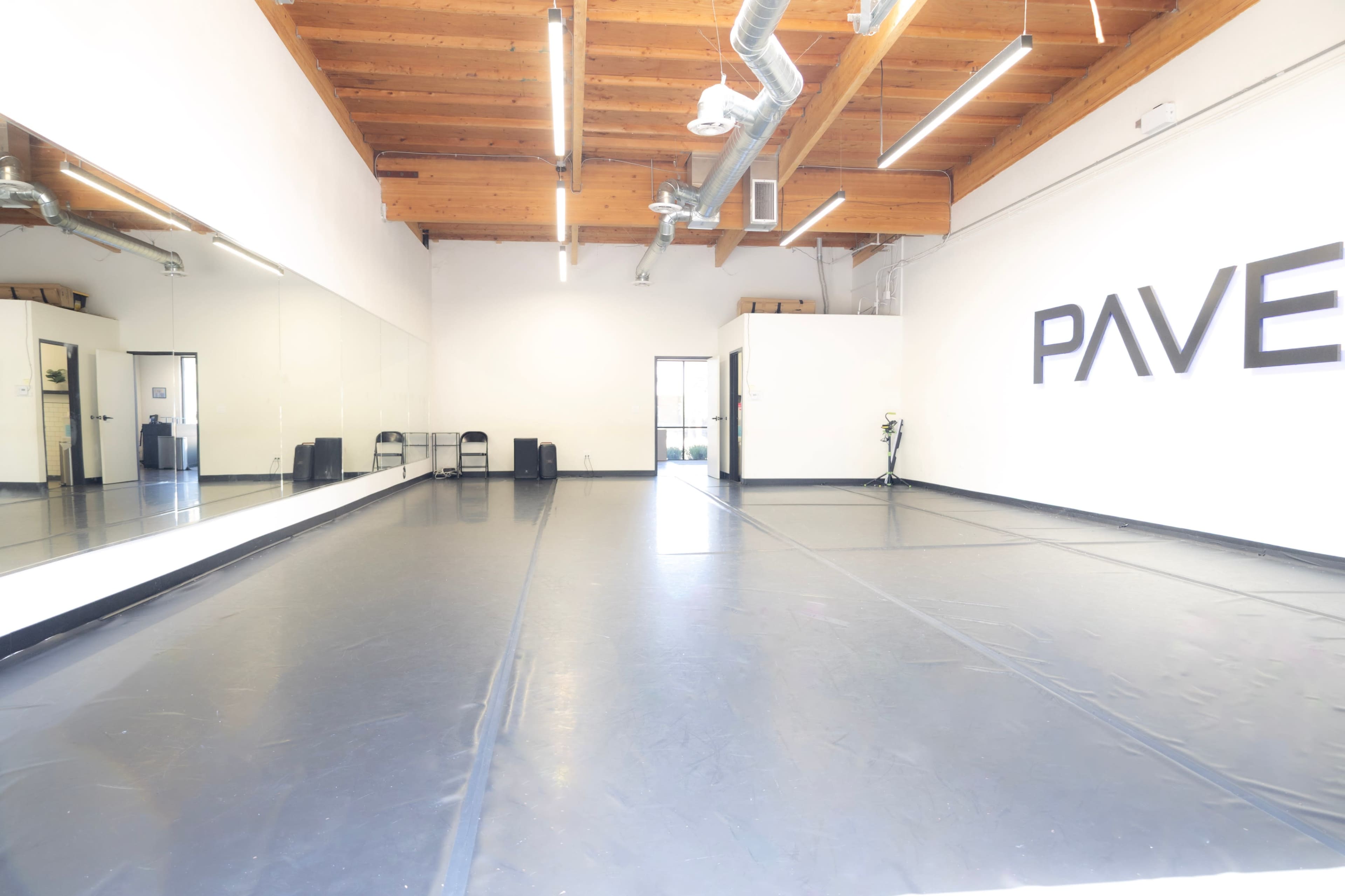 The image features a spacious dance studio with a black floor, mirrored wall, wooden ceiling beams, and the word "PAVE" displayed prominently on one wall.