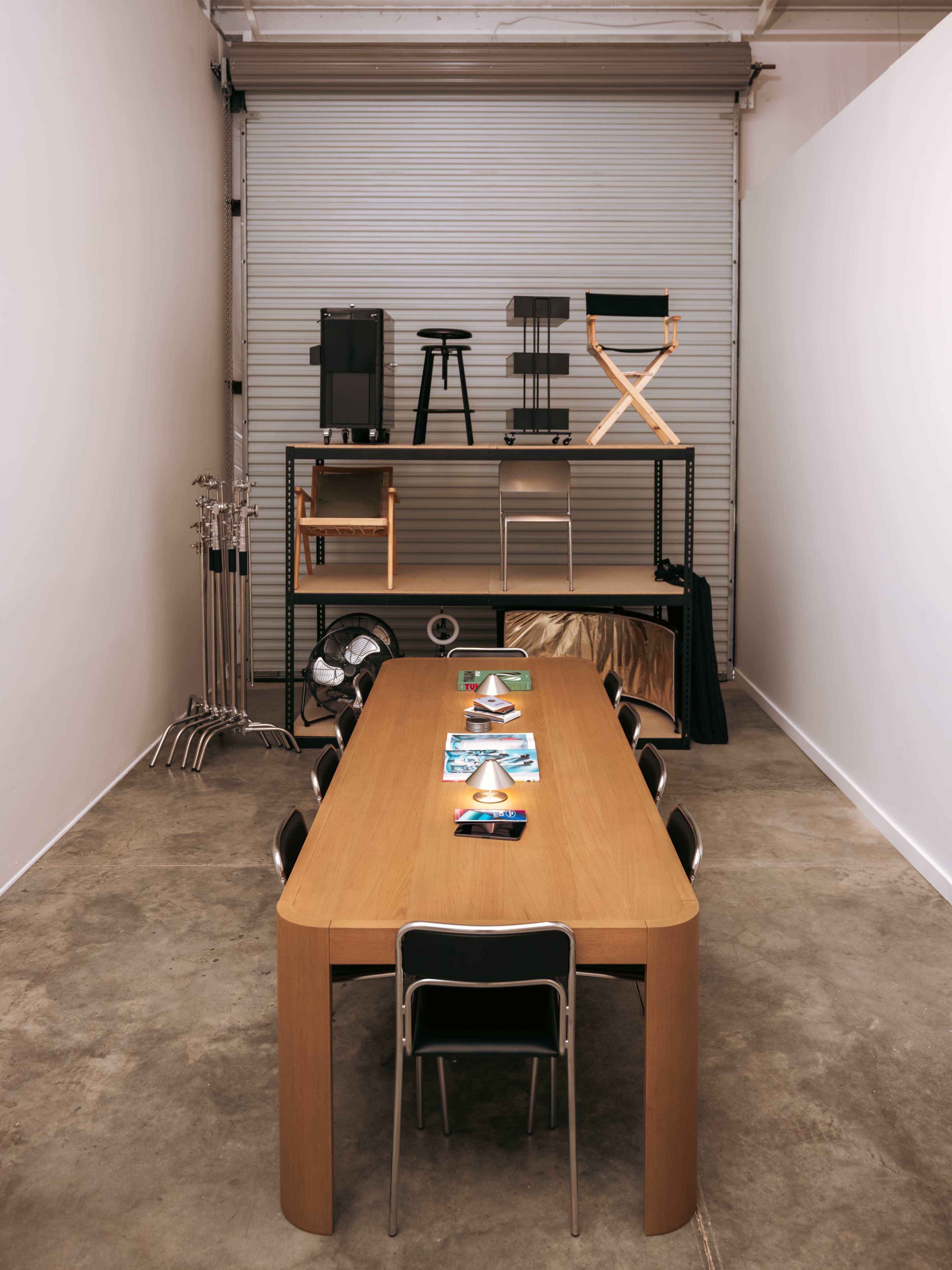 A long wooden table with black chairs is set in a minimalist room, featuring shelves above displaying various furniture pieces.