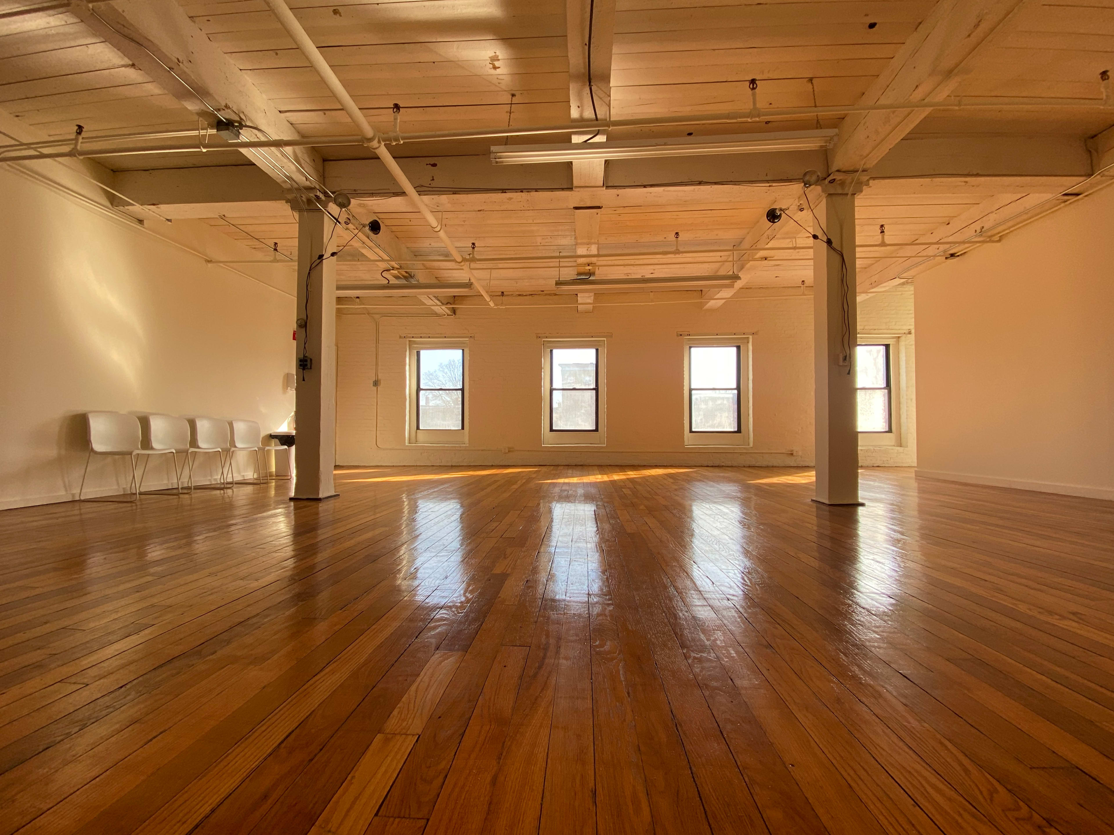 A spacious, sunlit room with polished wooden floors and five white chairs lined up against a wall, featuring large windows that let in natural light.