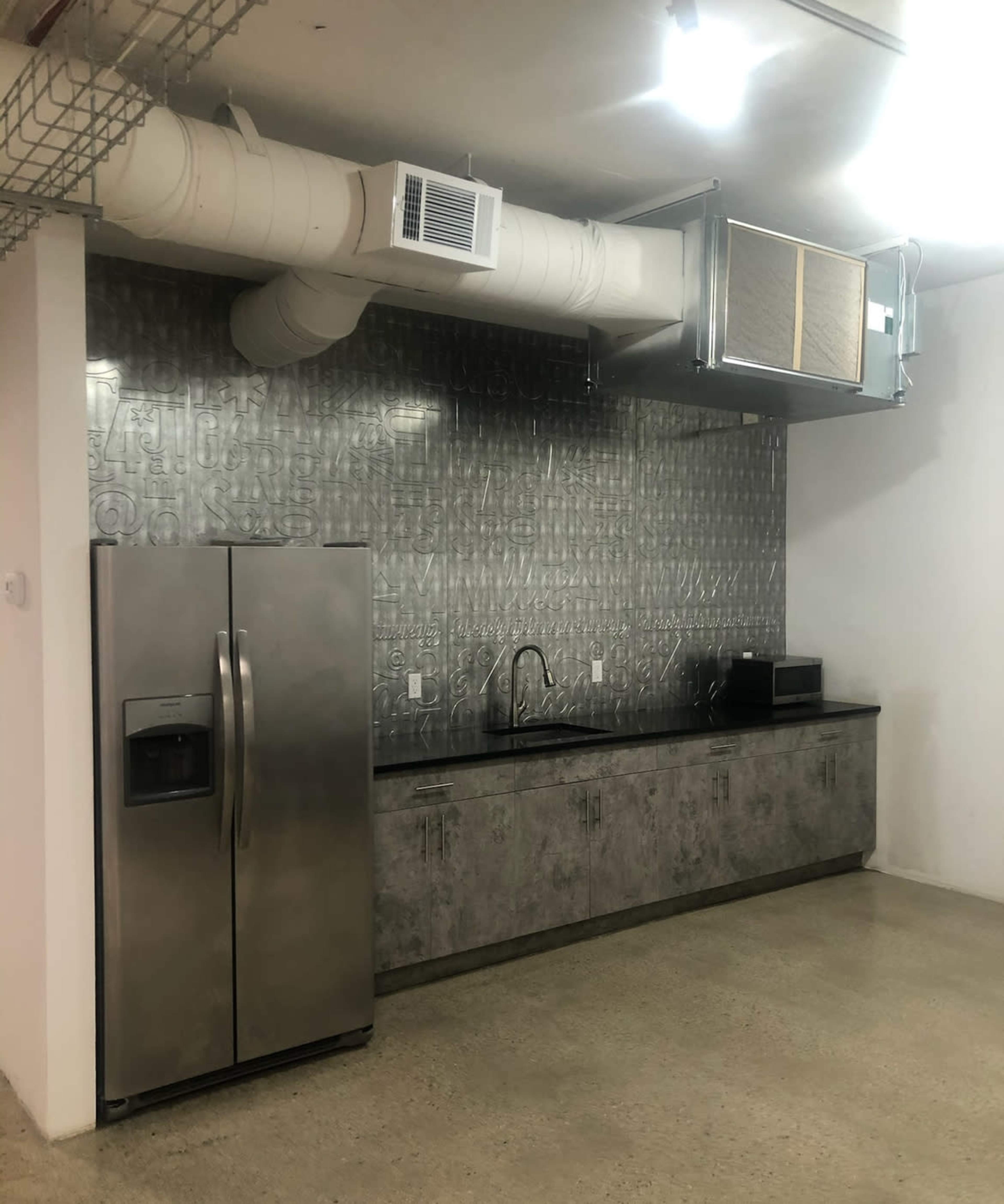 The image shows a modern kitchen with stainless steel appliances, including a refrigerator and a microwave, set against a textured metal backsplash and concrete cabinets.