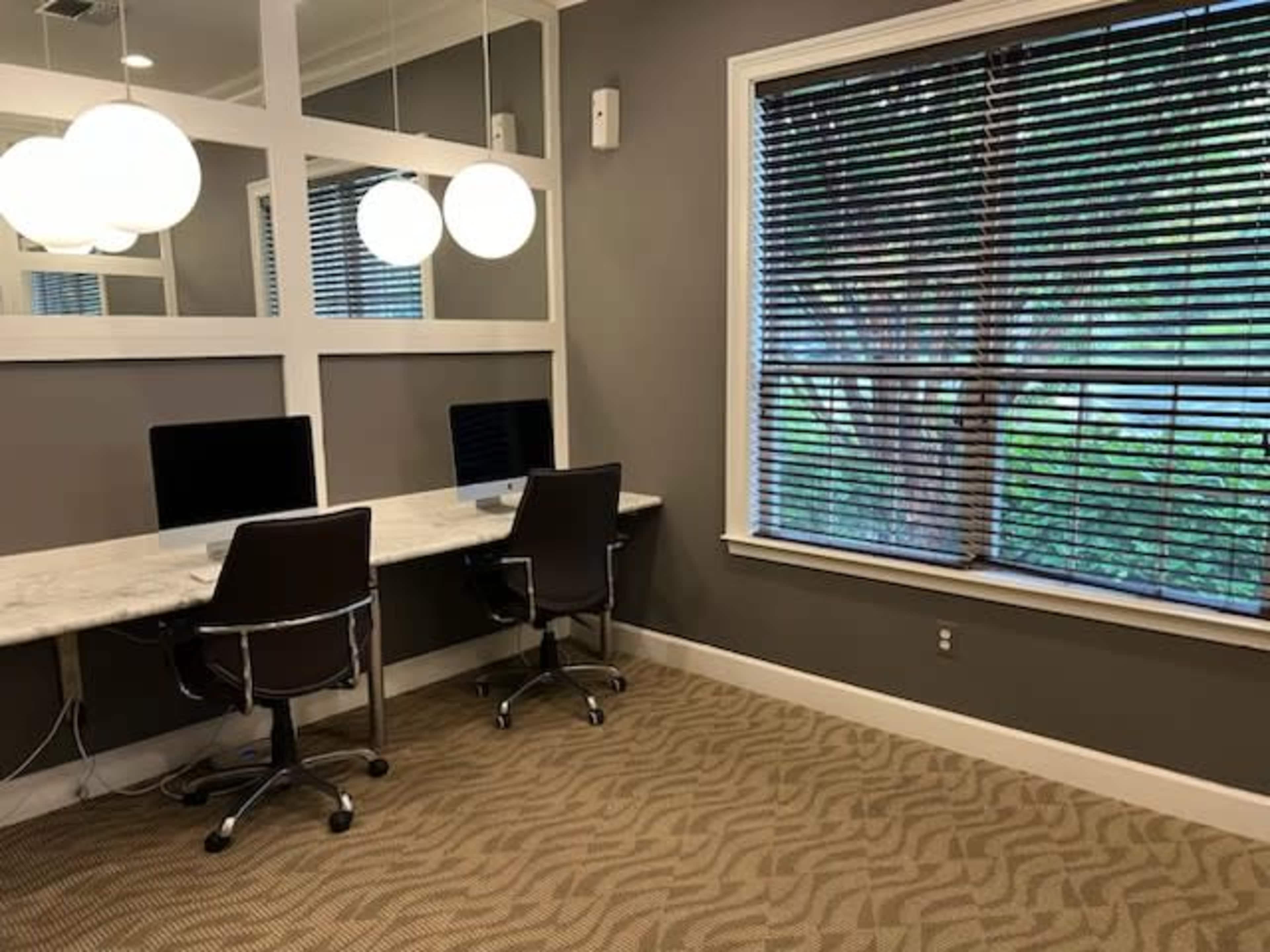 A small office space with two computer desks facing a window adorned with blinds and two round overhead lights.