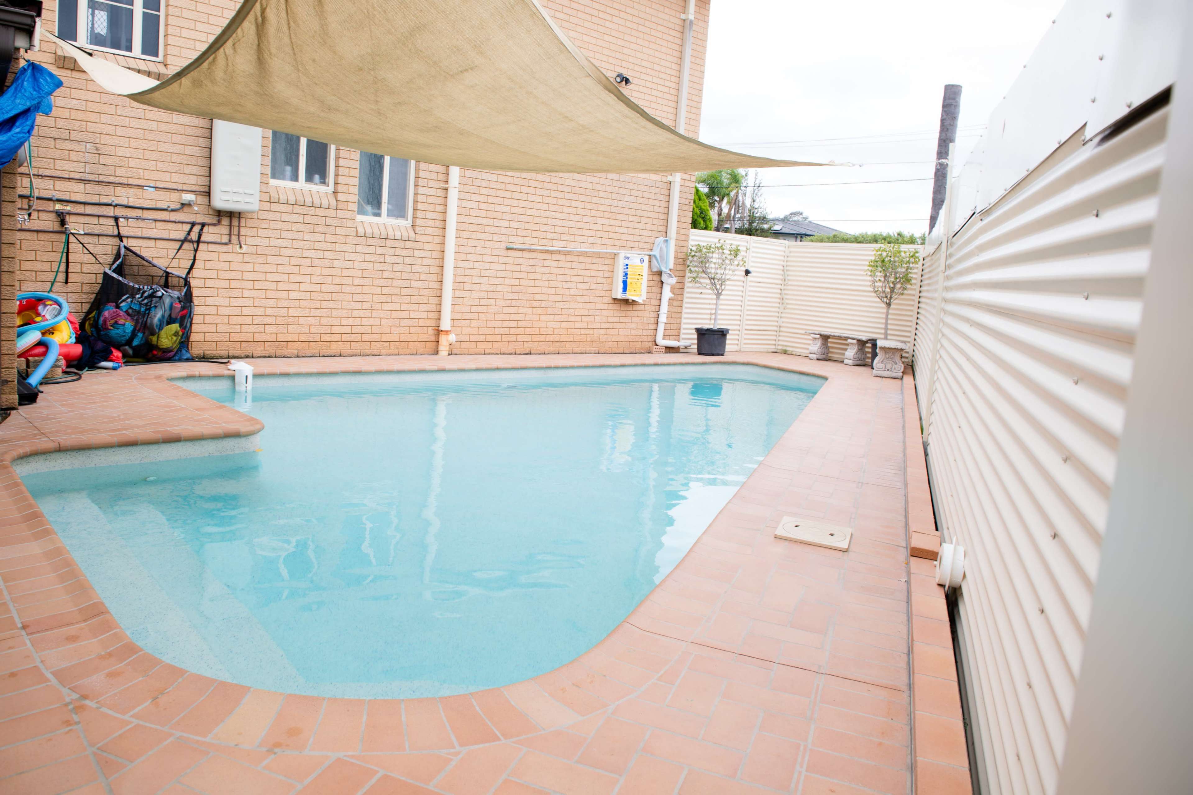 A rectangular swimming pool is bordered by brick tiles and features a shade sail overhead in a backyard setting.