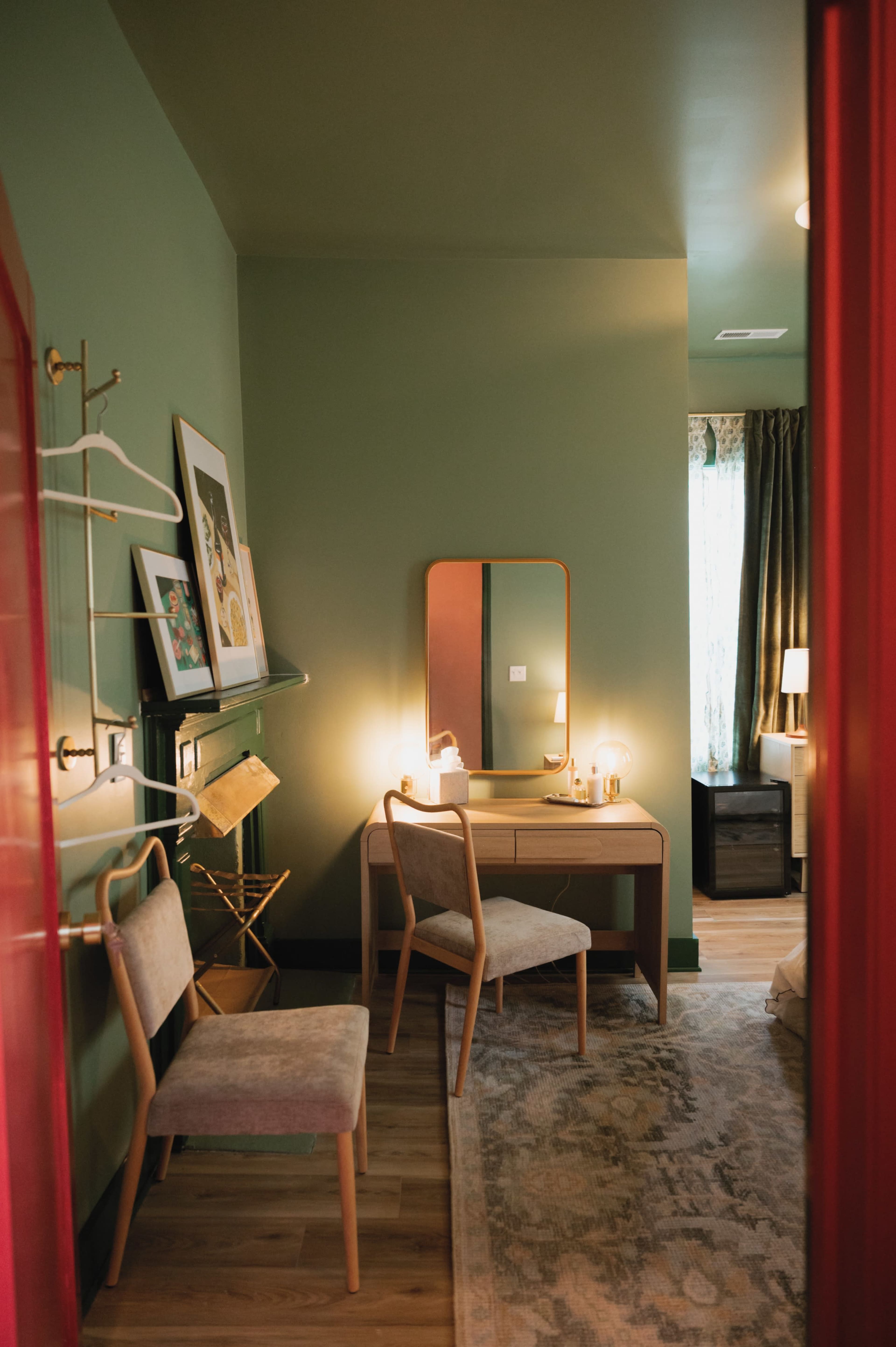 The image shows a small room with green walls, featuring a wooden desk with a mirror, two chairs, and framed artwork on the wall.