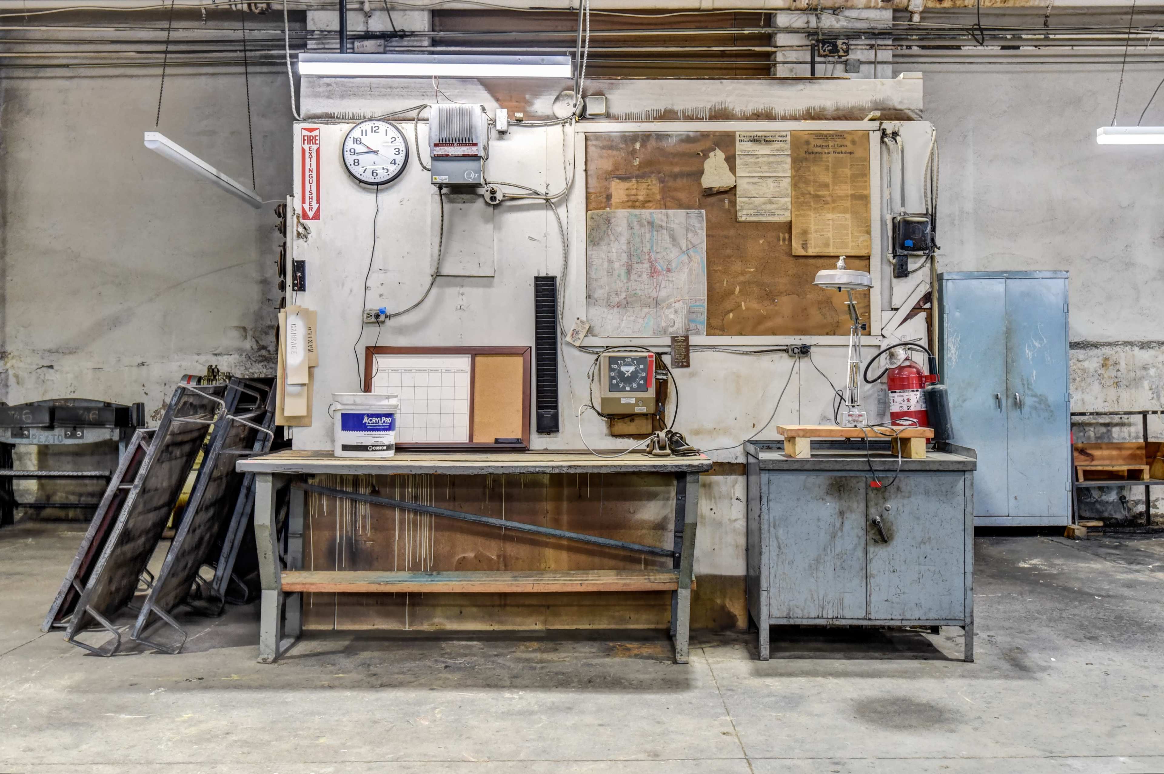 A cluttered workshop with a large workbench, various tools, a clock, and a metal cabinet against a bare wall.