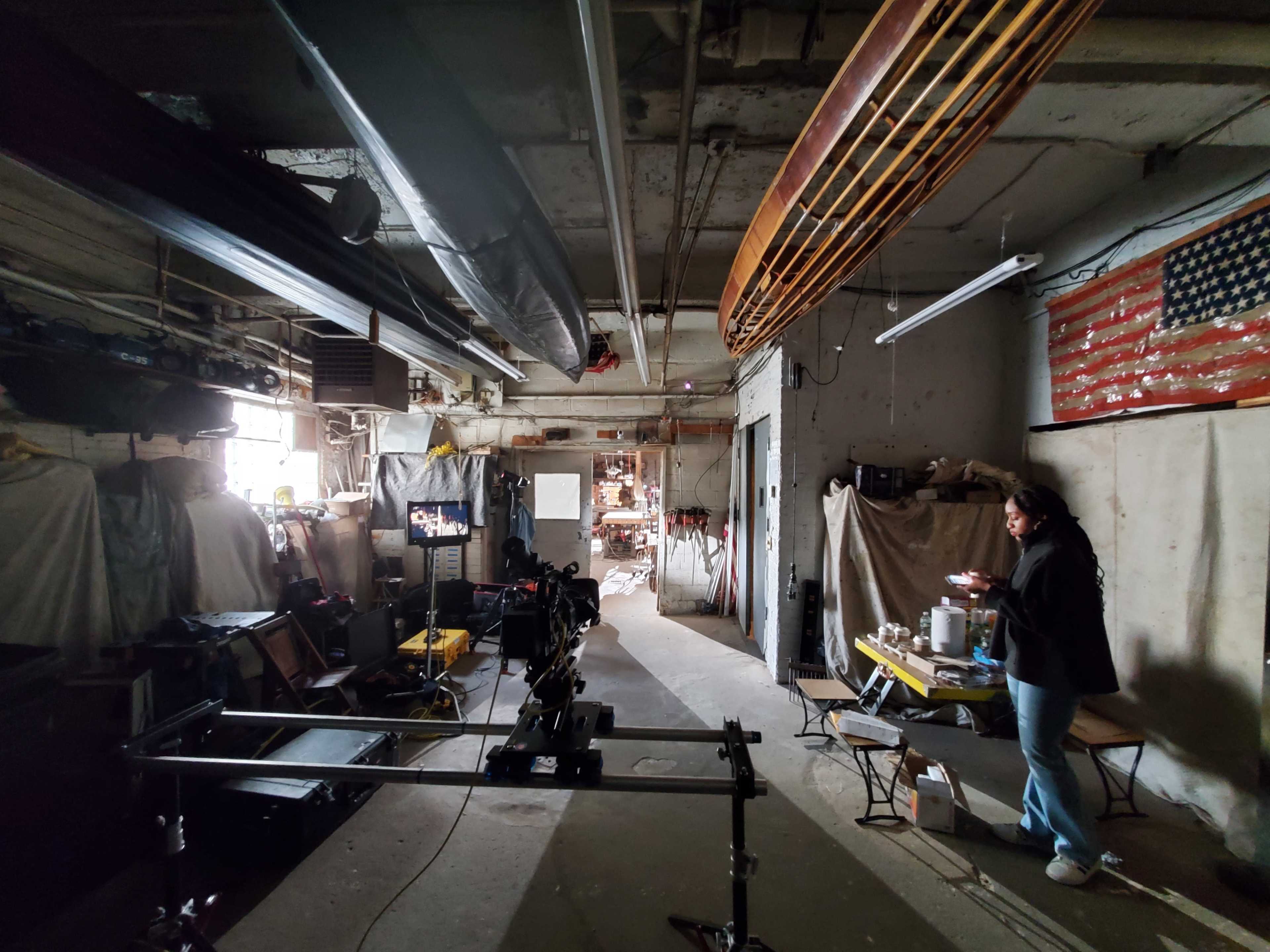 A woman stands by the wall in a dimly lit workshop, surrounded by various tools and materials, with a partially visible American flag in the background.
