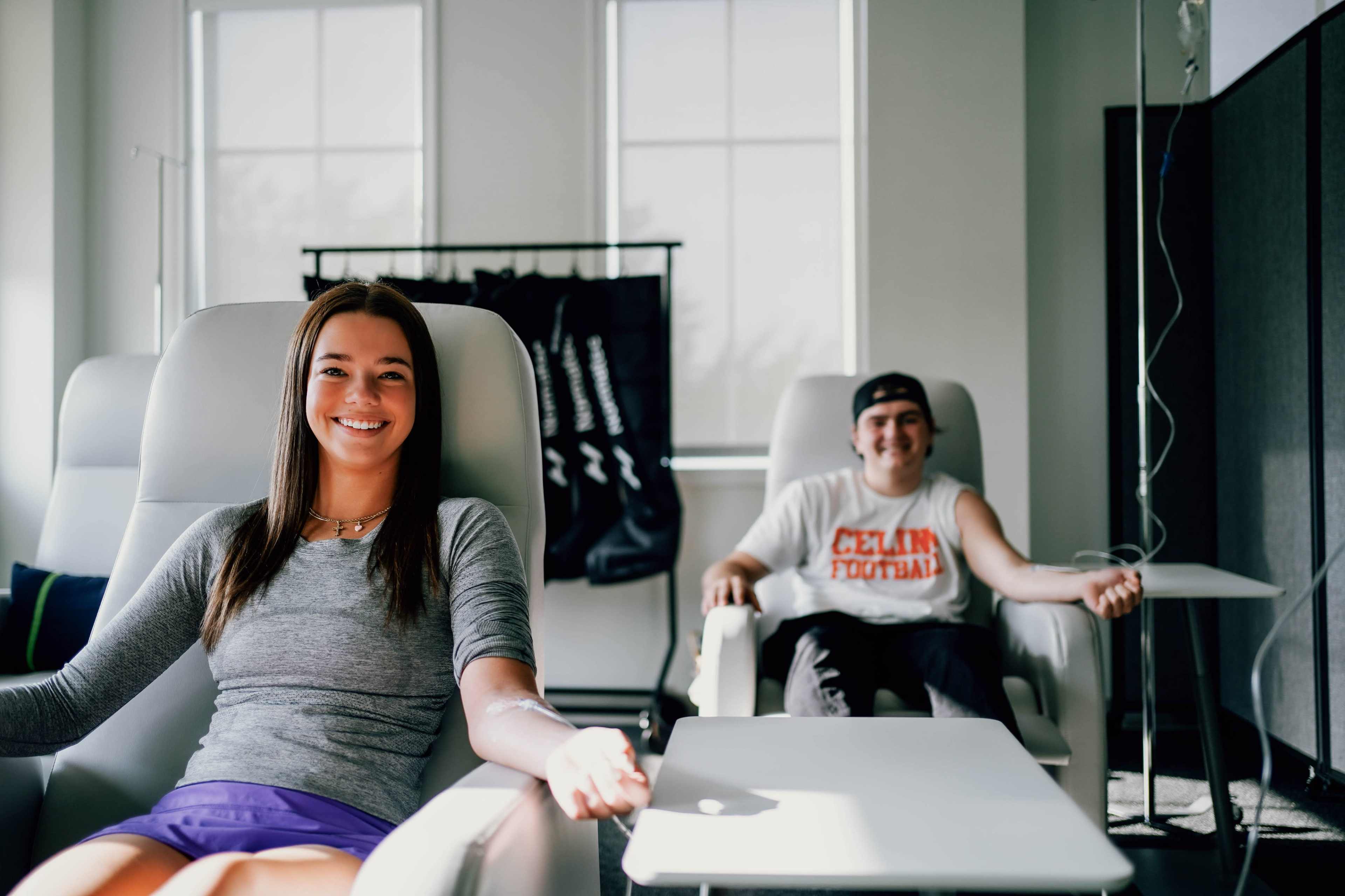 Two individuals sit in modern recliners in a bright room, each connected to an IV while a football-themed garment hangs in the background.