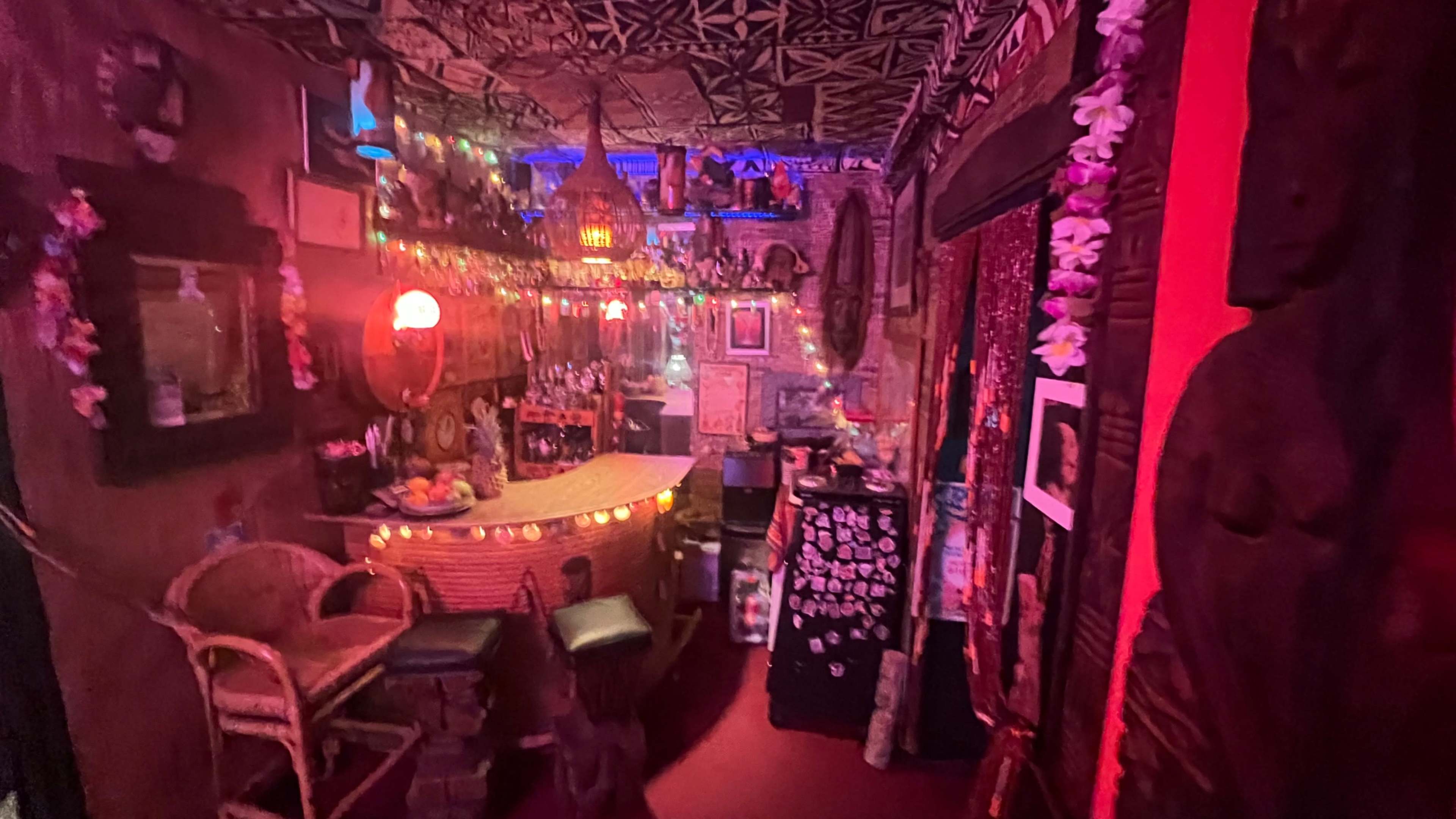 The image shows a dimly lit bar area decorated with colorful string lights, tropical decor, and various artworks lining the walls.