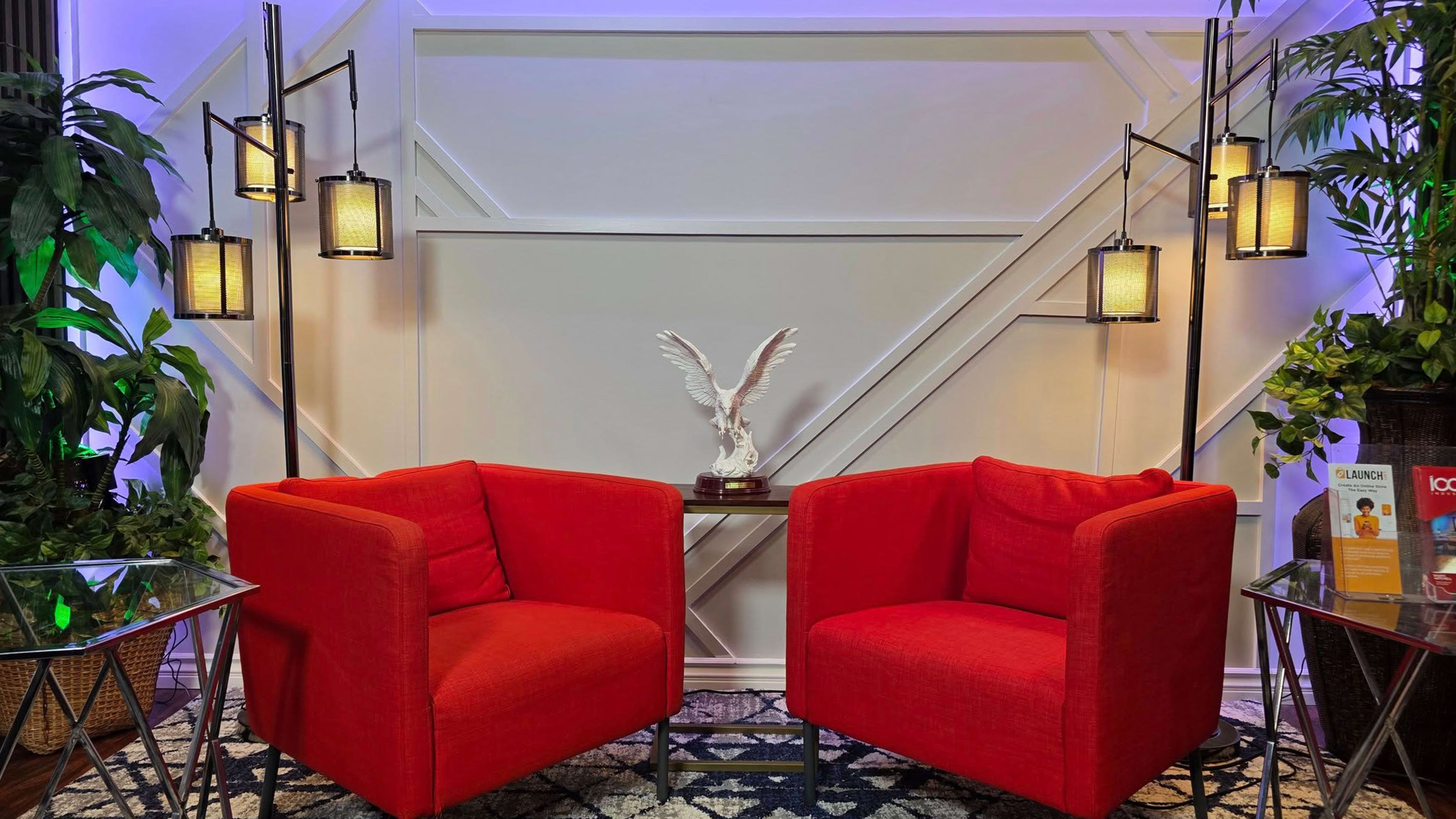 The image shows a cozy seating area with two red armchairs positioned around a small table, flanked by plants and decorative lighting.