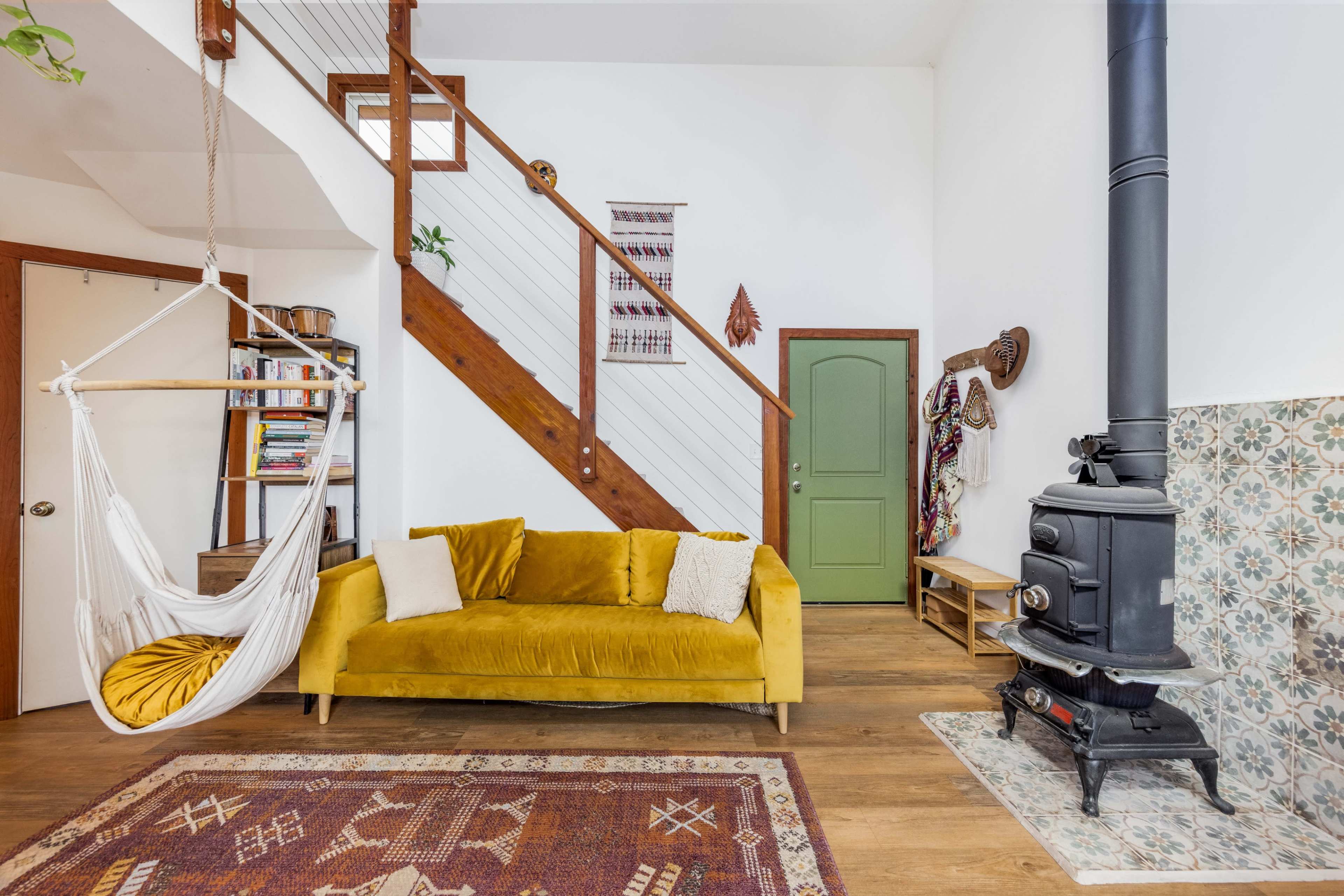 A cozy living room with a yellow couch, a hanging chair, a staircase, and a black wood-burning stove beside decorative tiles.
