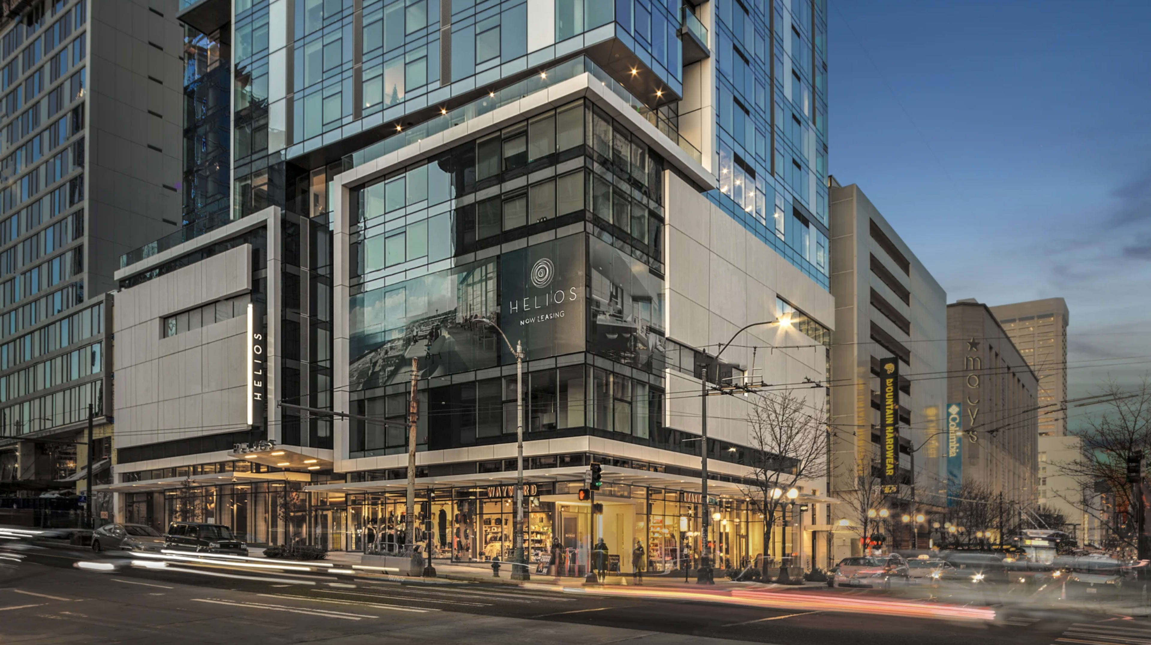 The image shows a modern multi-story building with large glass windows and a corner storefront, located at a busy urban intersection during dusk.