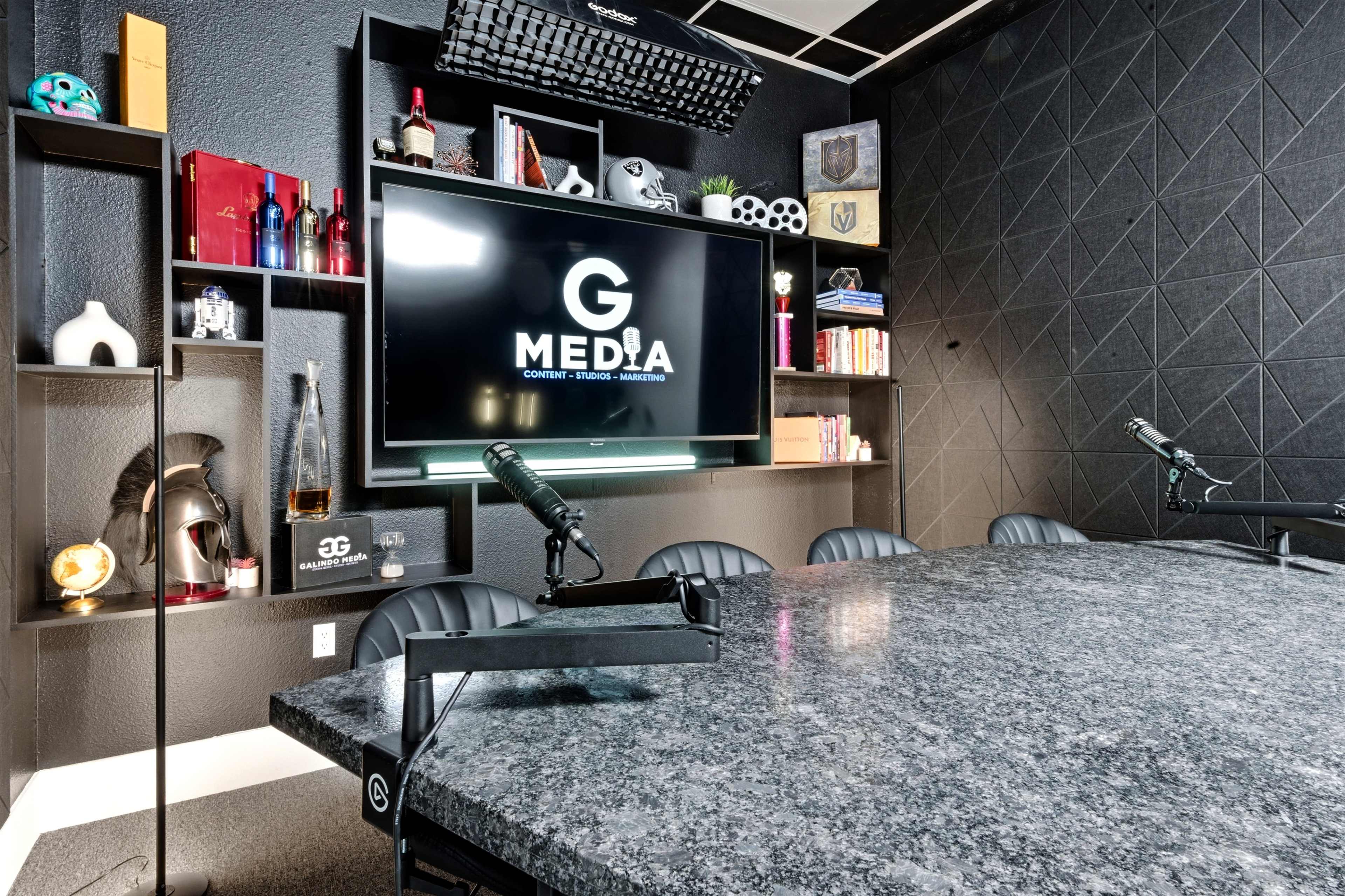 The image shows a modern media office featuring a large granite table, a wall-mounted screen displaying the logo "G MEDIA," and various decorative items on shelves.