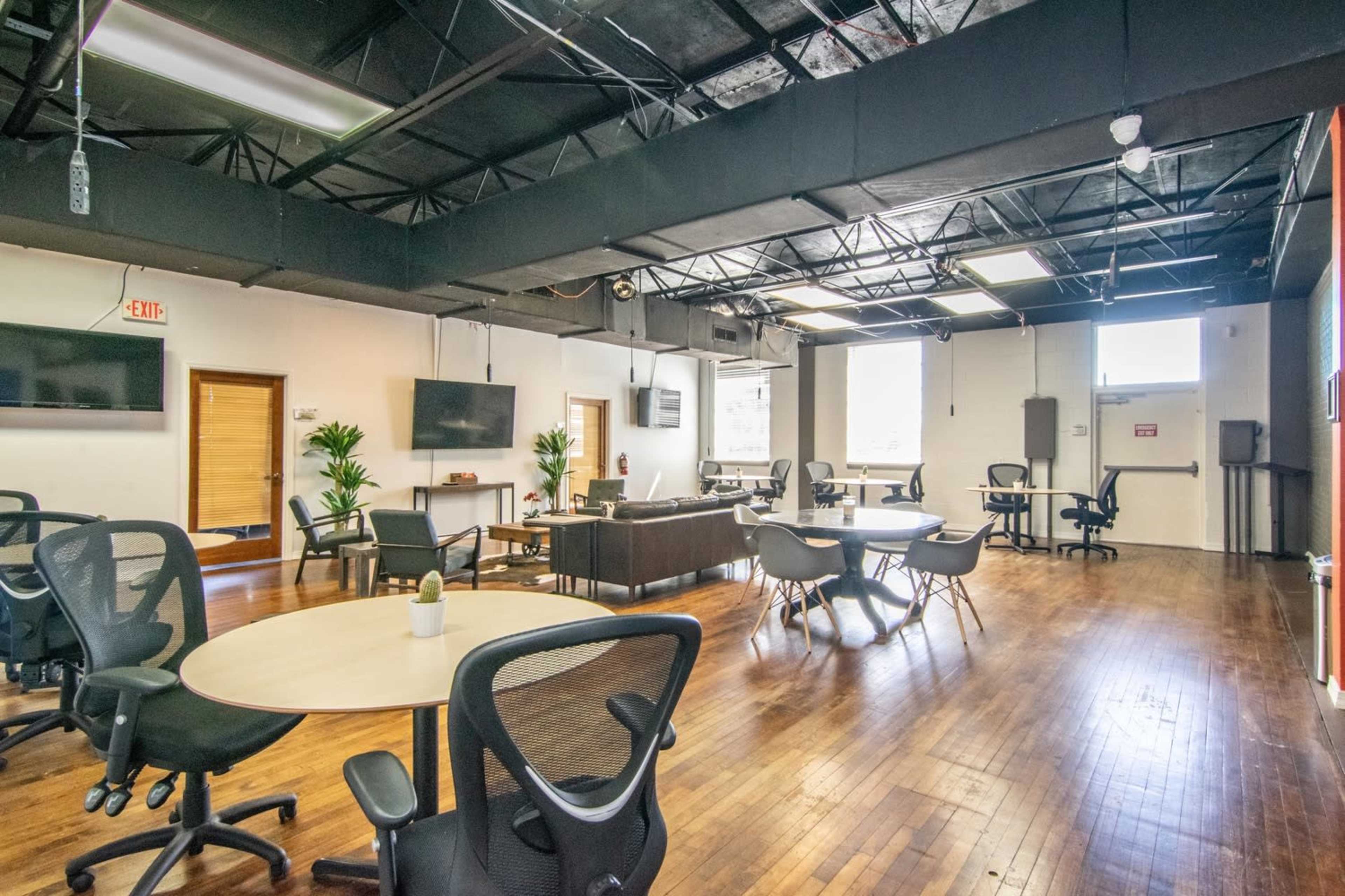 A spacious coworking area with multiple tables, chairs, and plants, featuring large windows that allow natural light to enter.