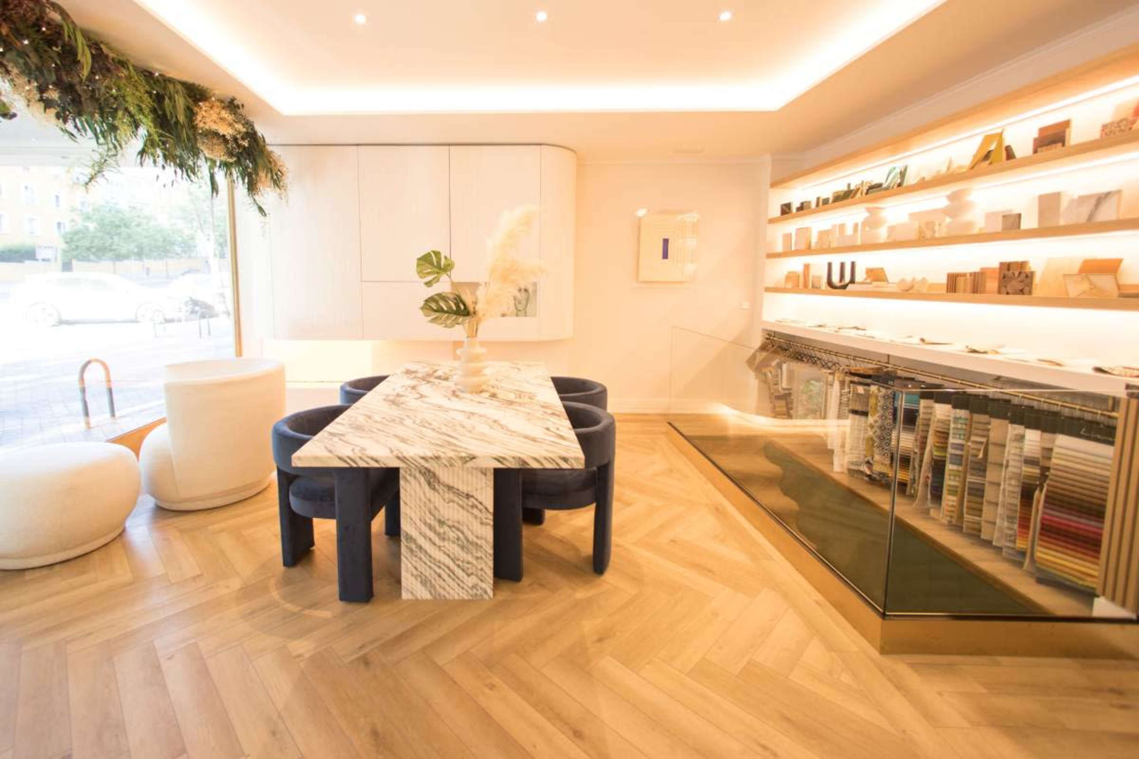 The image shows a modern interior space featuring a marble dining table surrounded by navy blue chairs, with shelves displaying various items and warm lighting.