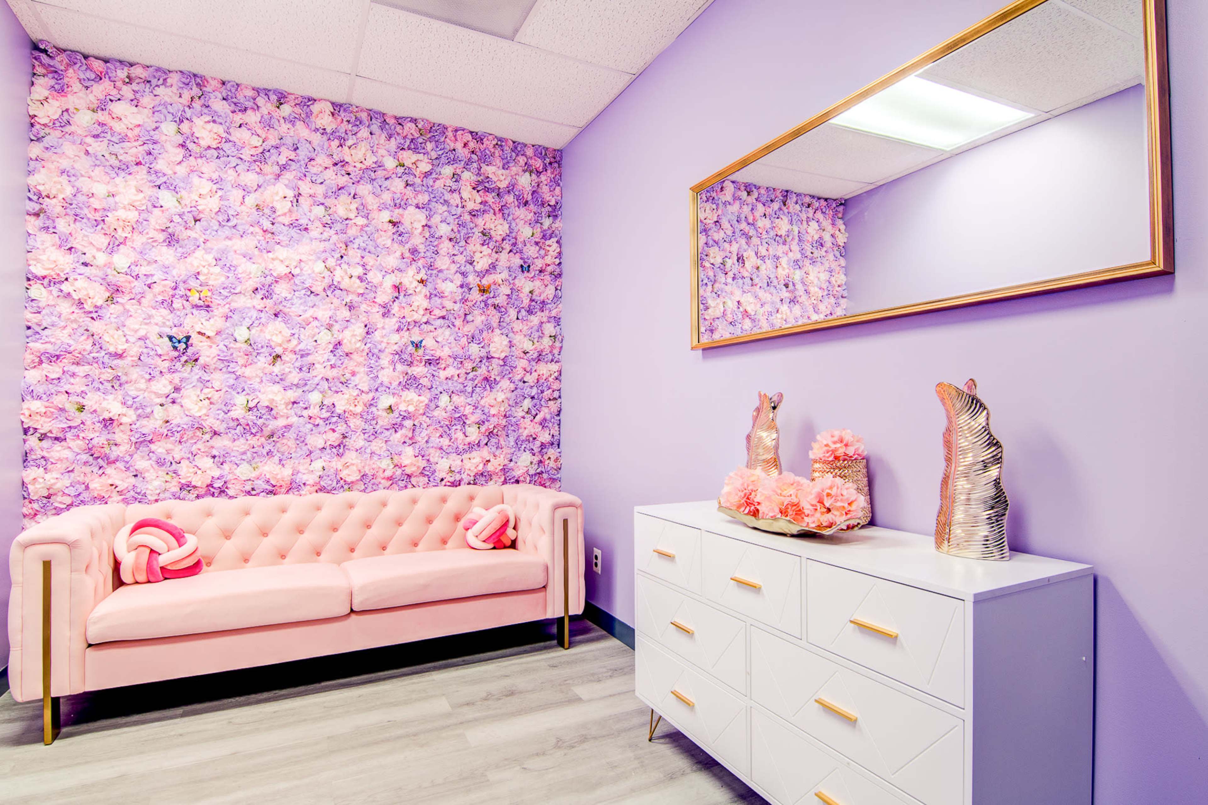 A styled room featuring a pink tufted sofa, a white dresser with decorative items, and a flower-patterned purple accent wall.