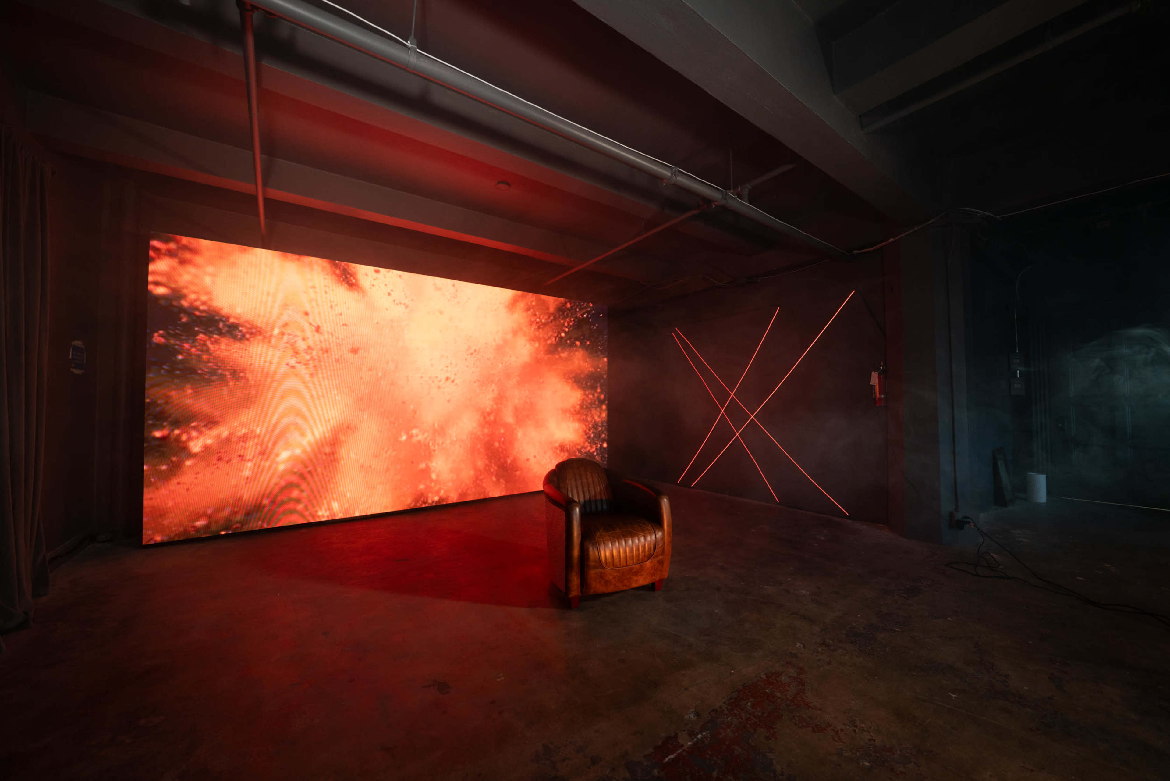 The image shows a room with a brown leather chair placed in front of a large, bright screen displaying fiery visuals, and a wall featuring red neon light patterns.