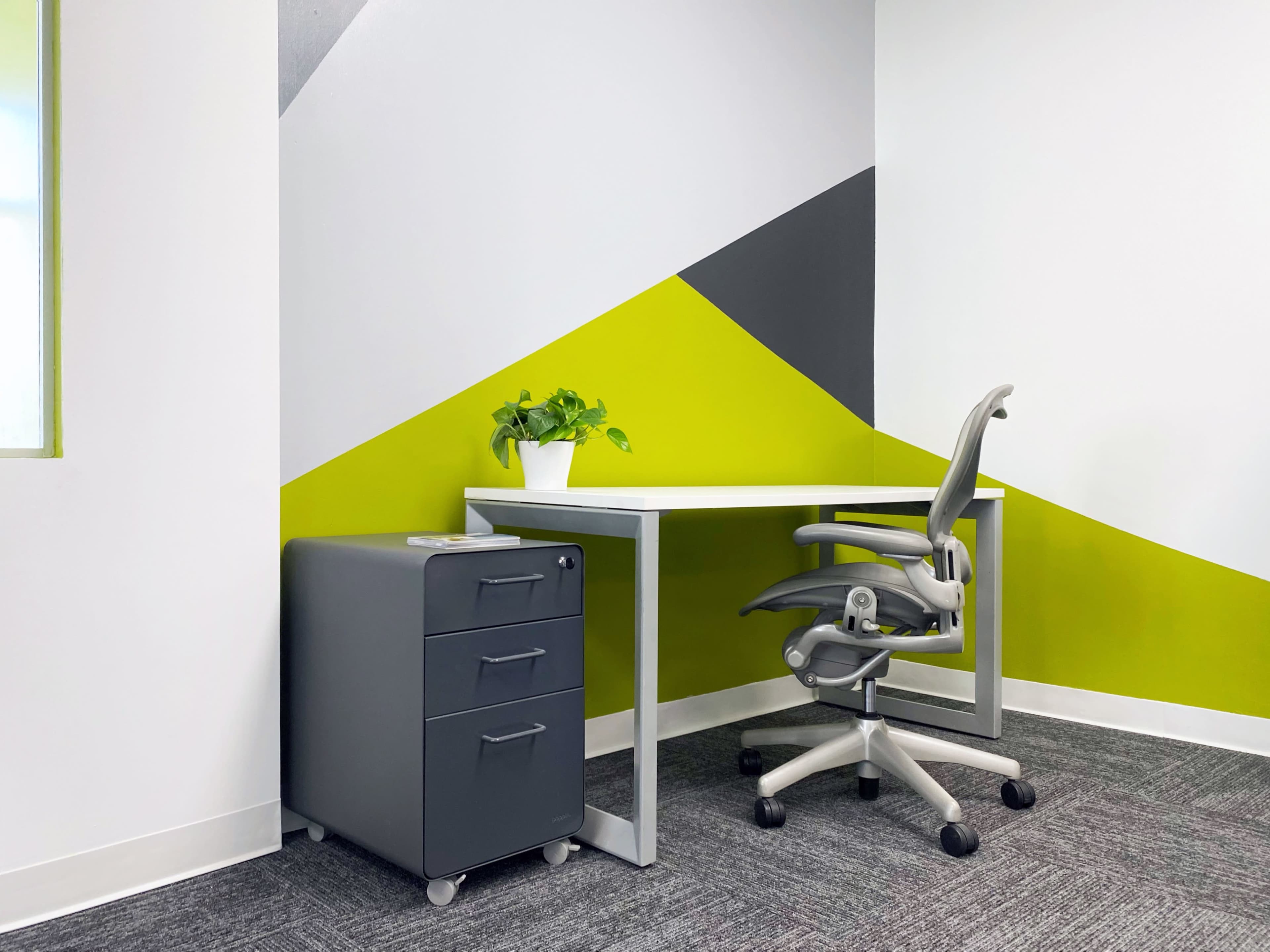 A modern office space with a white desk, an ergonomic chair, and a gray rolling storage unit next to a brightly colored geometric wall.