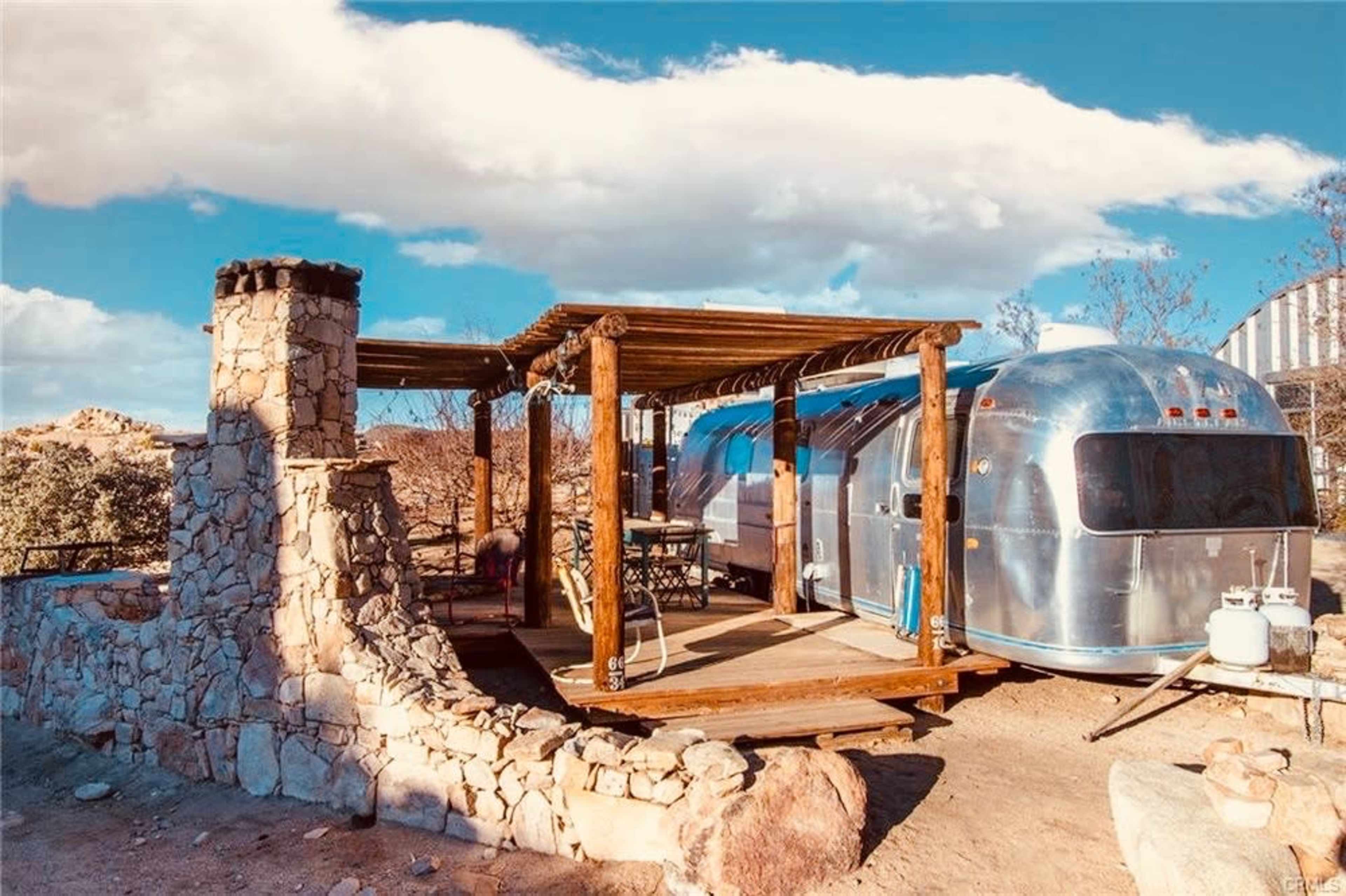 A vintage Airstream trailer is parked beside a stone structure with a wooden pergola in a desert landscape under a blue sky.