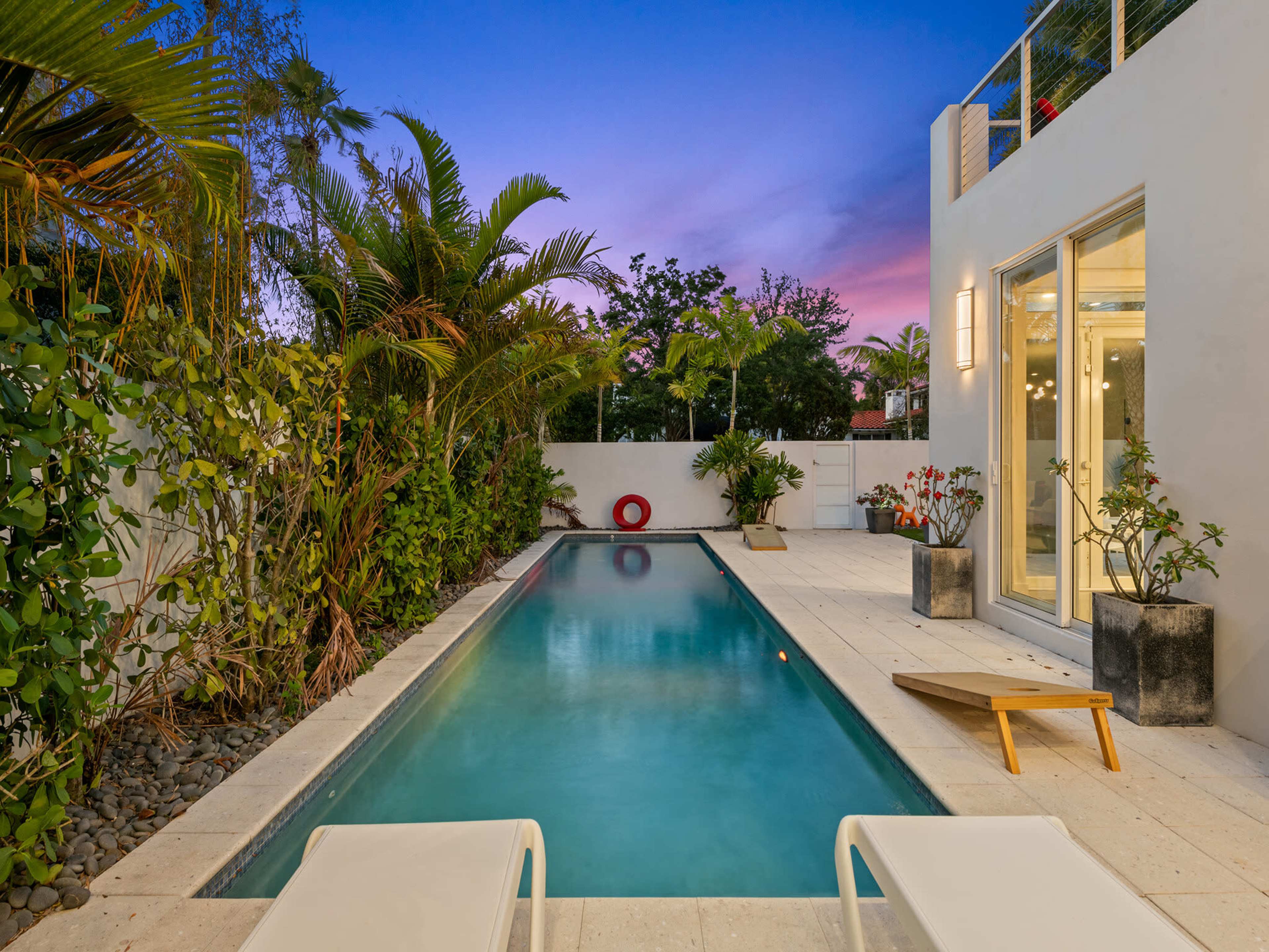 The image shows a modern outdoor pool area surrounded by lush greenery and a colorful sunset sky.