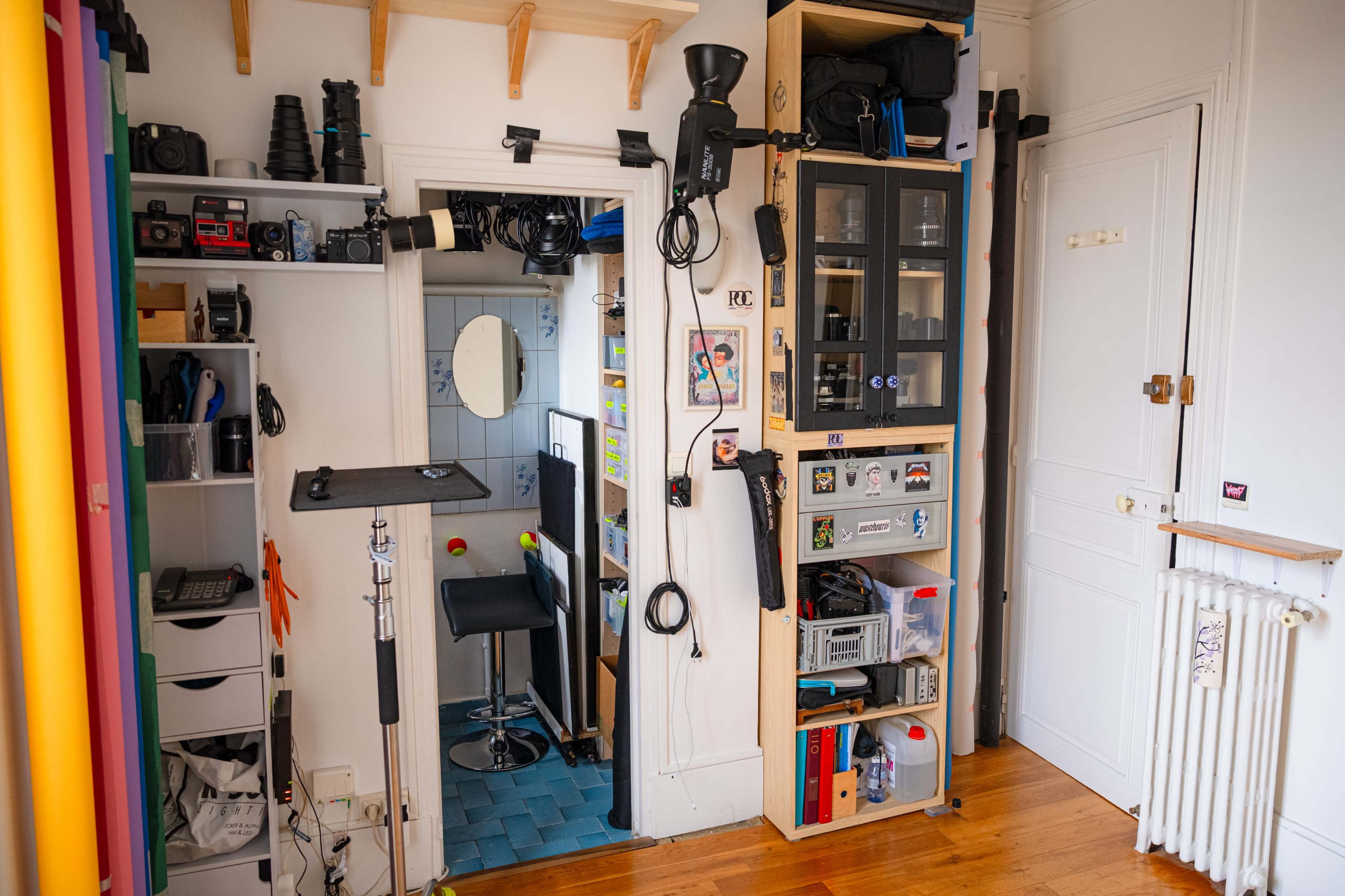 The image shows a room with various photography equipment and accessories organized on shelves, with a doorway leading to another area that appears to contain additional gear and a mirror.