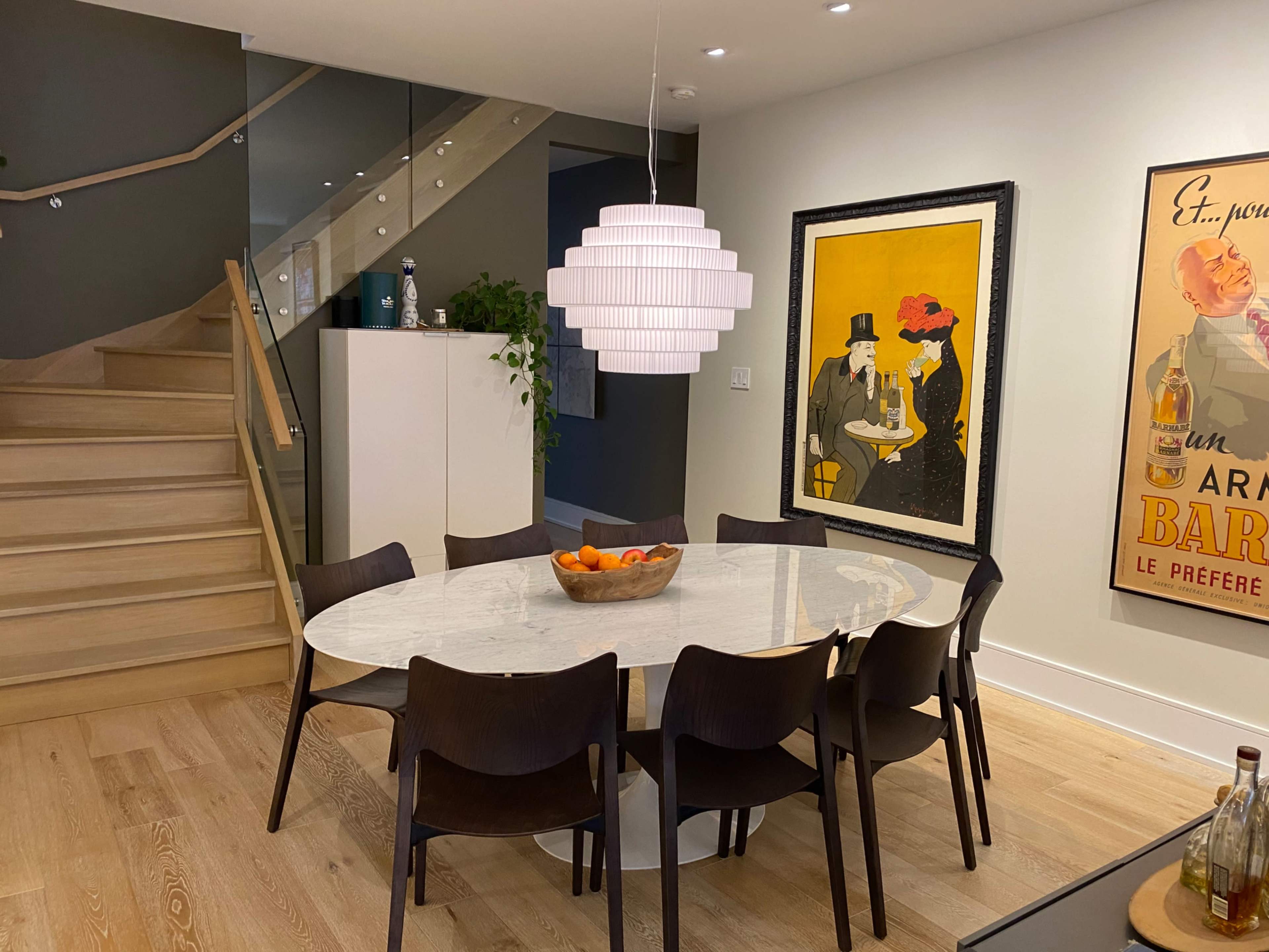 A round dining table with eight chairs is set in a modern room featuring a staircase, a pendant light, and framed artwork on the walls.