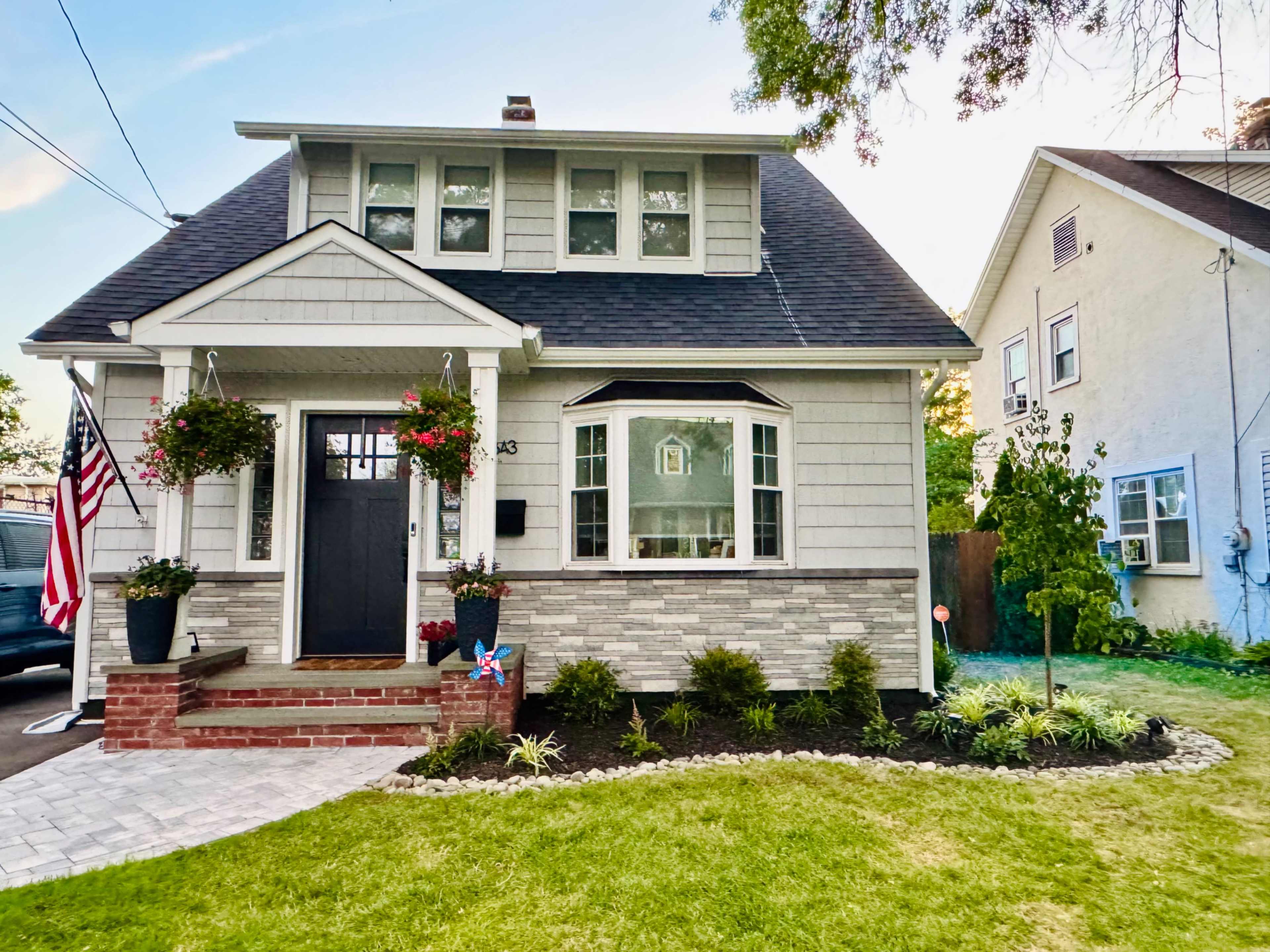 The image shows a two-story house with a gray exterior, a front porch, hanging flower baskets, and a neatly landscaped yard.