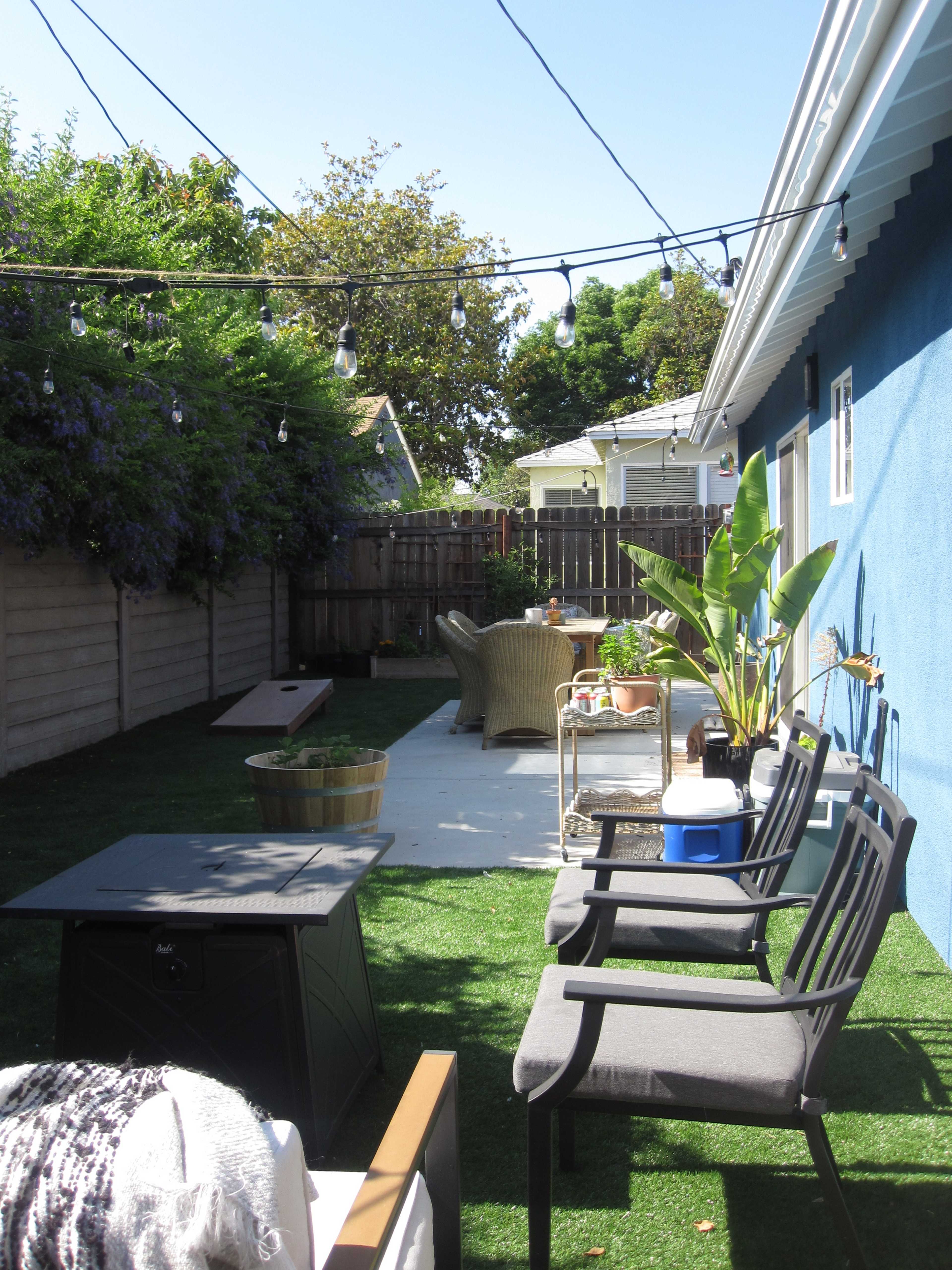 The image shows a backyard patio with artificial grass, seating arrangements, potted plants, and string lights overhead.