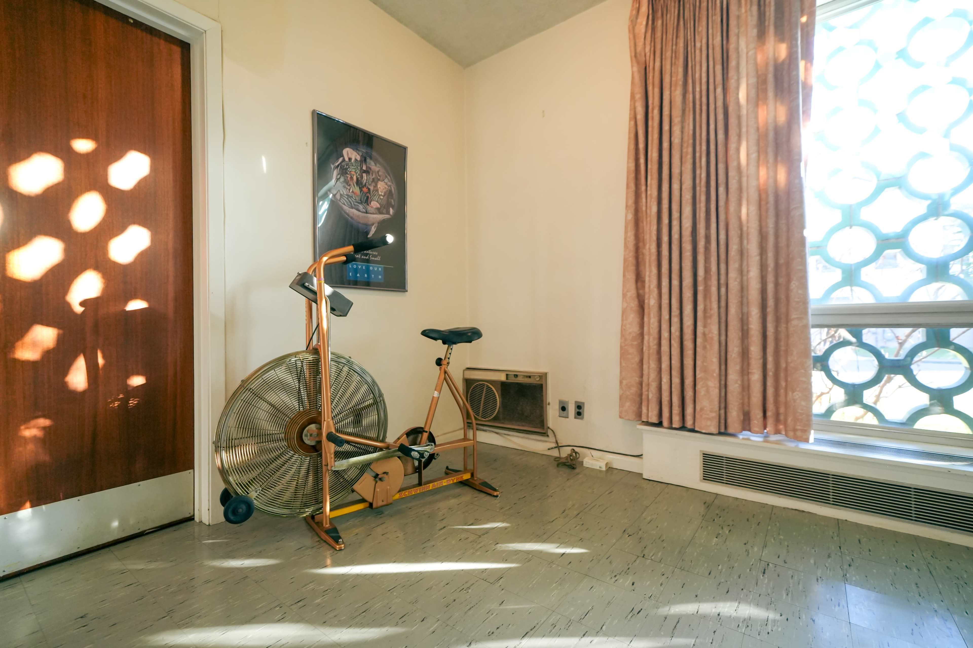 The image shows a room with a stationary exercise bike next to a wall-mounted air conditioning unit and a window covered by sheer curtains.