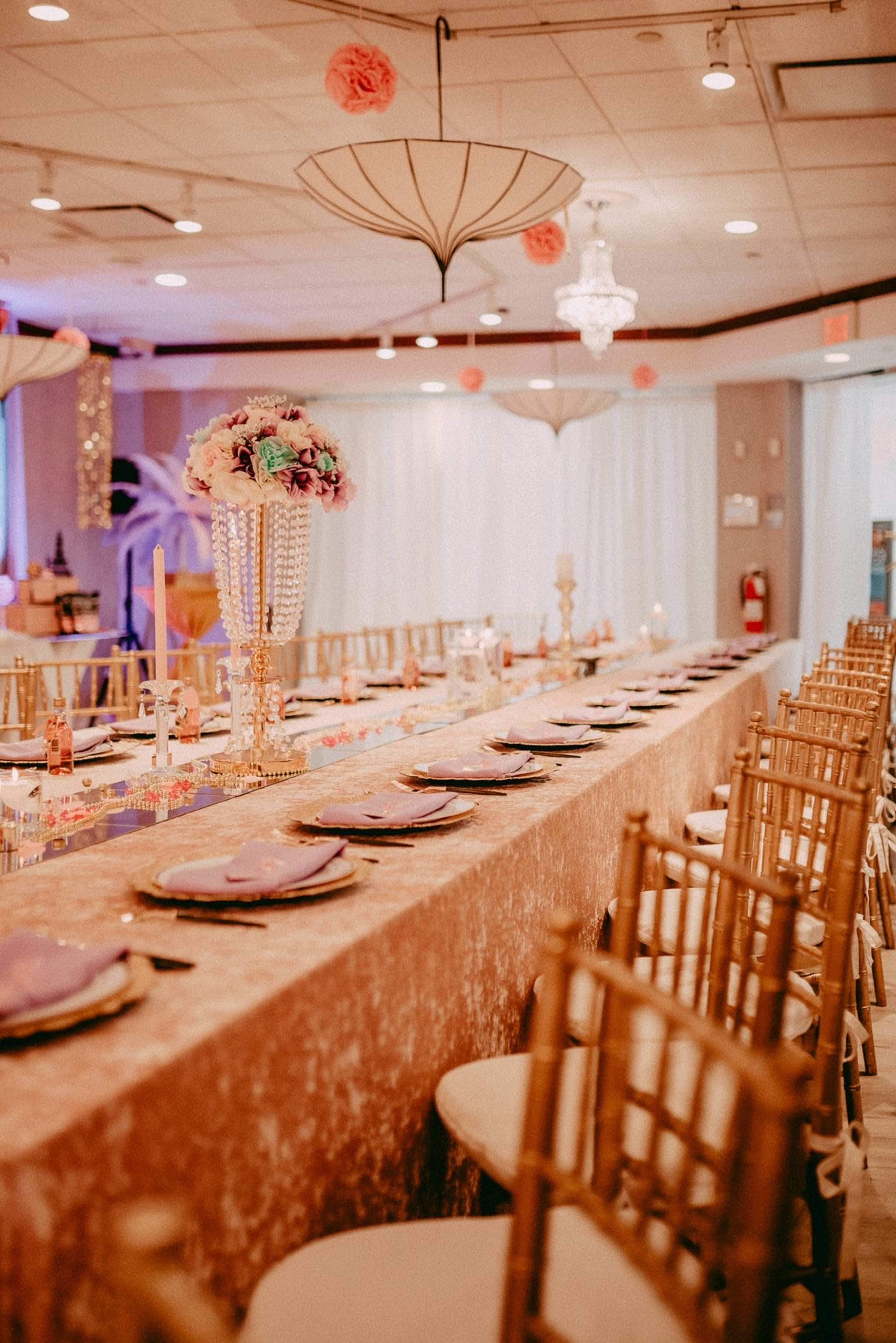 A long banquet table is set with elegant place settings, adorned with a decorative centerpiece and draped in a sparkling cloth under stylish lighting fixtures.