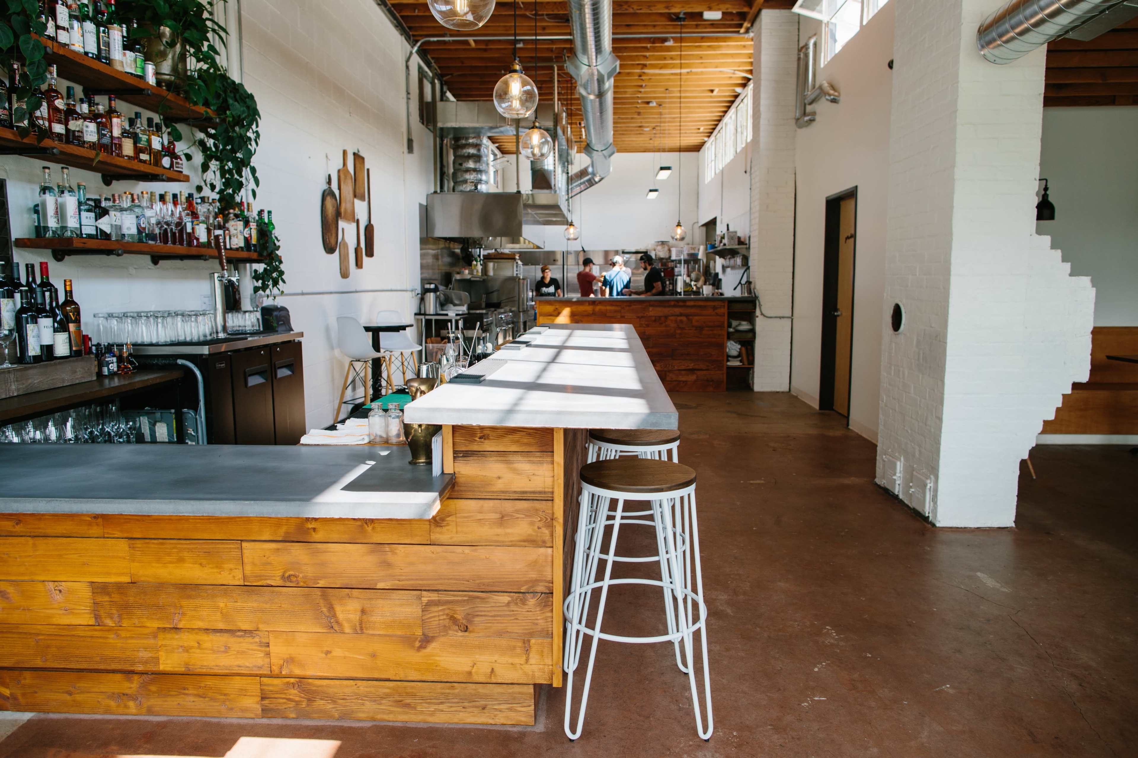 A spacious restaurant interior features a wooden bar with high stools, shelves holding various bottles, and a open kitchen area in the background.