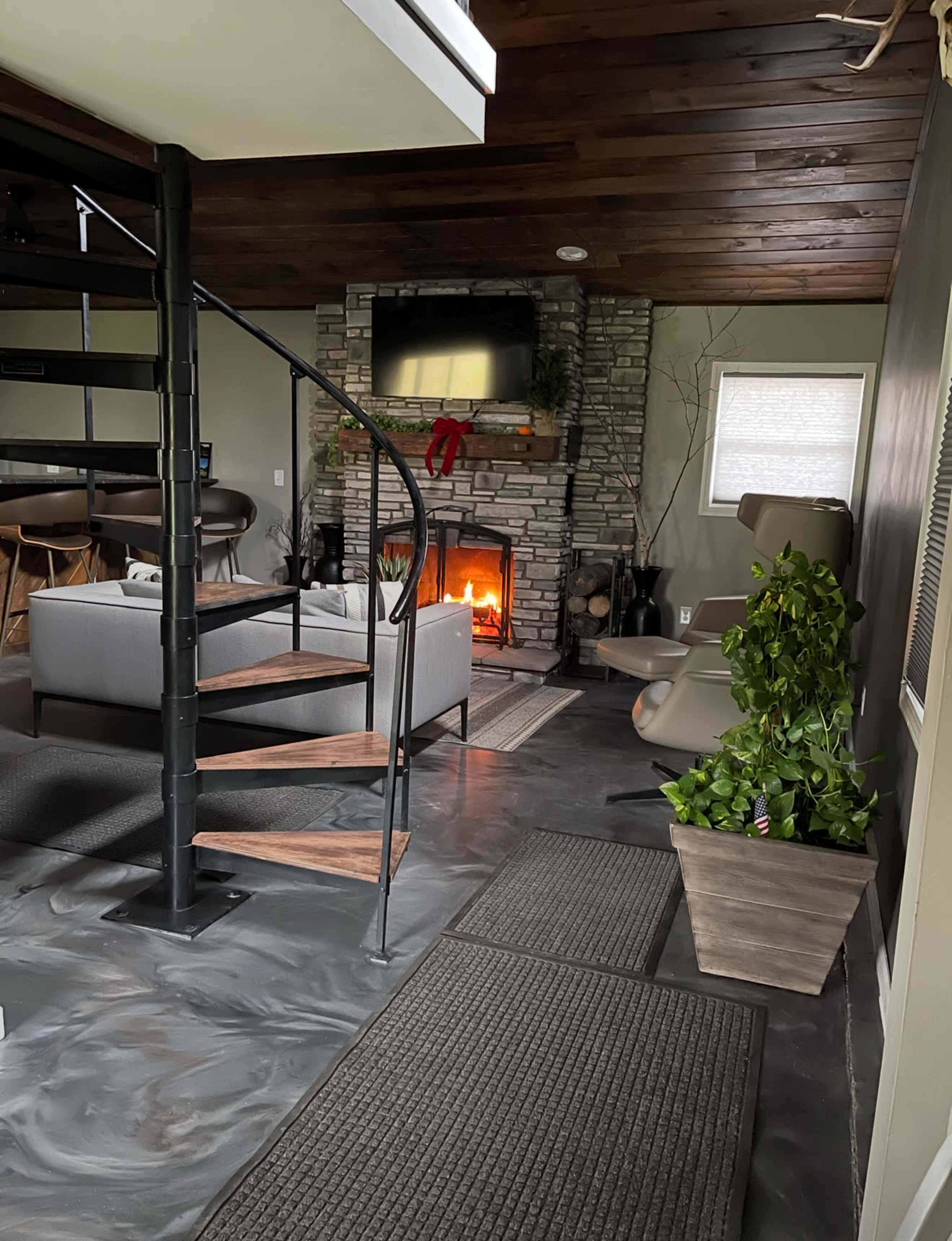 The image shows a modern living room featuring a spiral staircase, a stone fireplace with a fire burning, and contemporary furniture arranged around the fireplace.