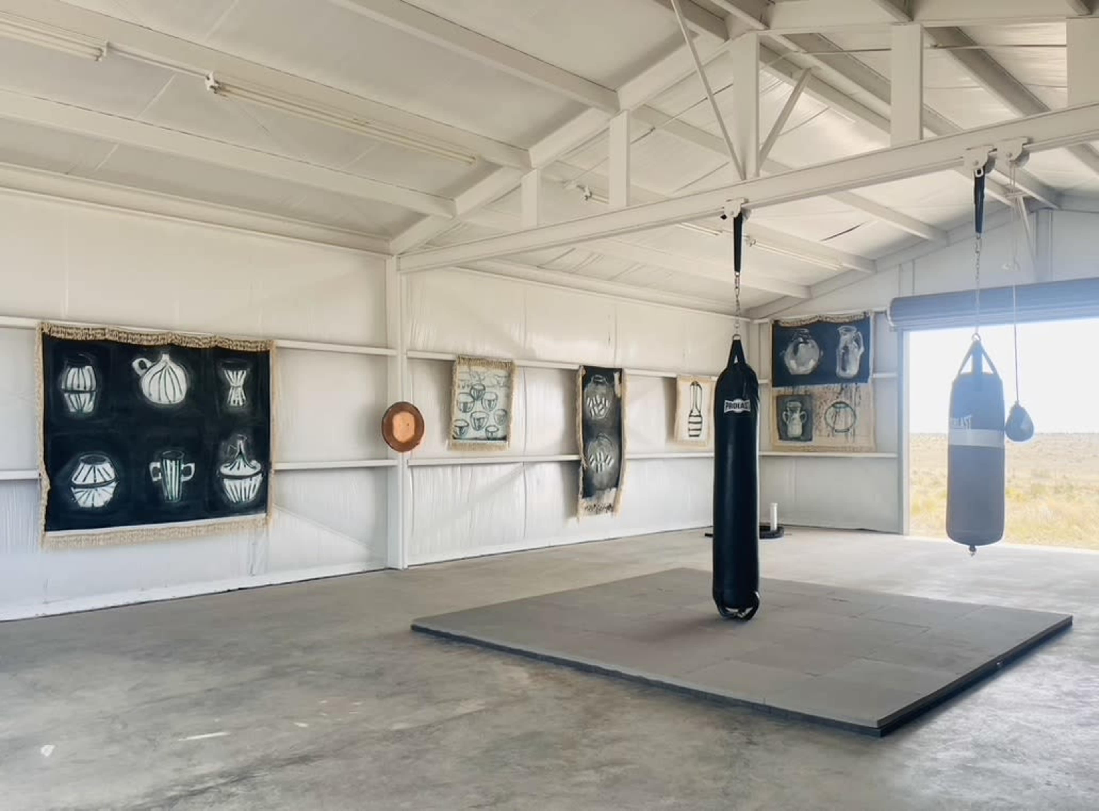 The image shows a spacious room with white walls, featuring a black punching bag suspended from the ceiling, surrounded by various artworks displayed on the walls.
