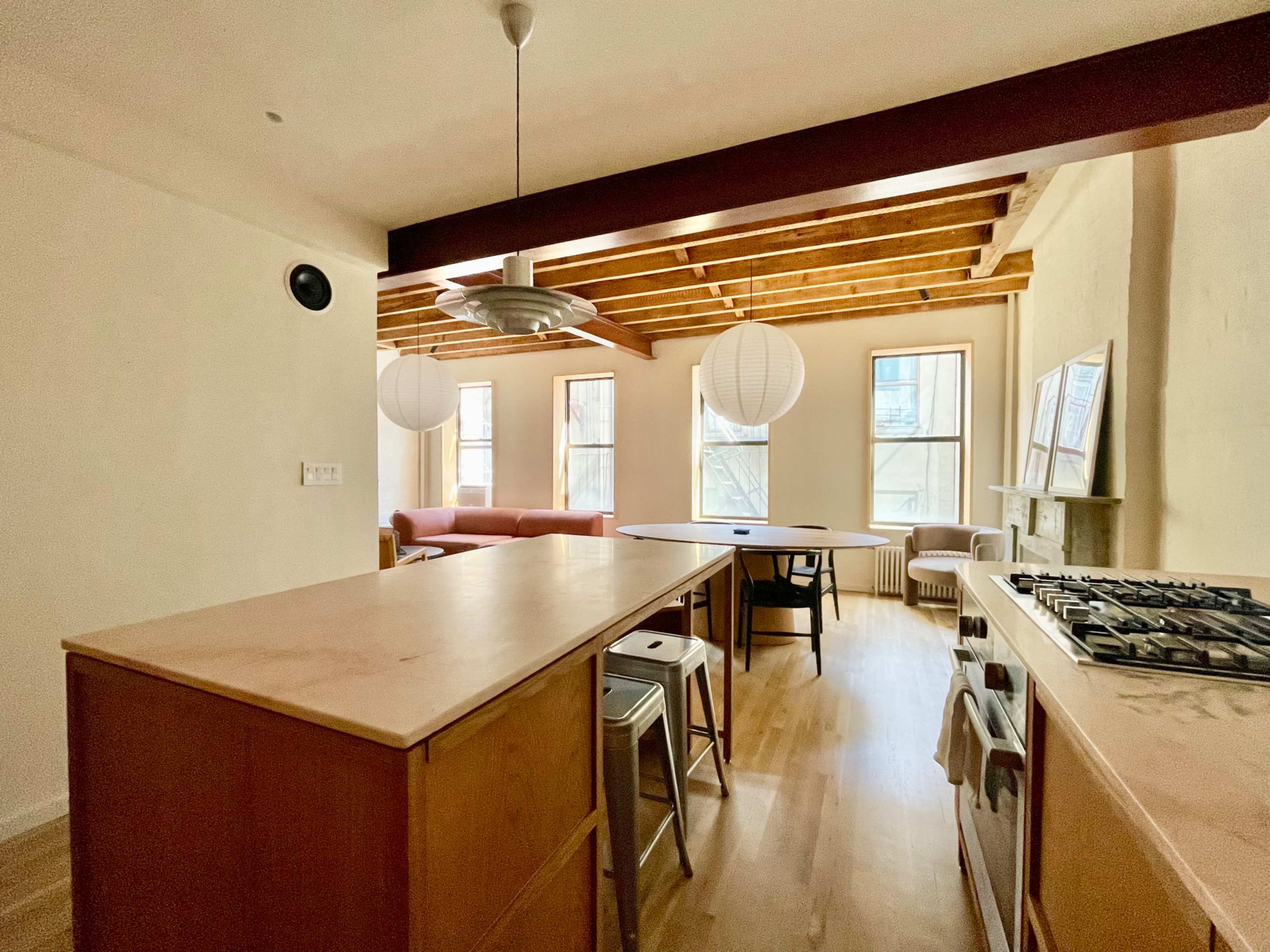The image shows a modern kitchen area with wooden beams, a central island, and a dining table, located adjacent to a living space with a sofa.