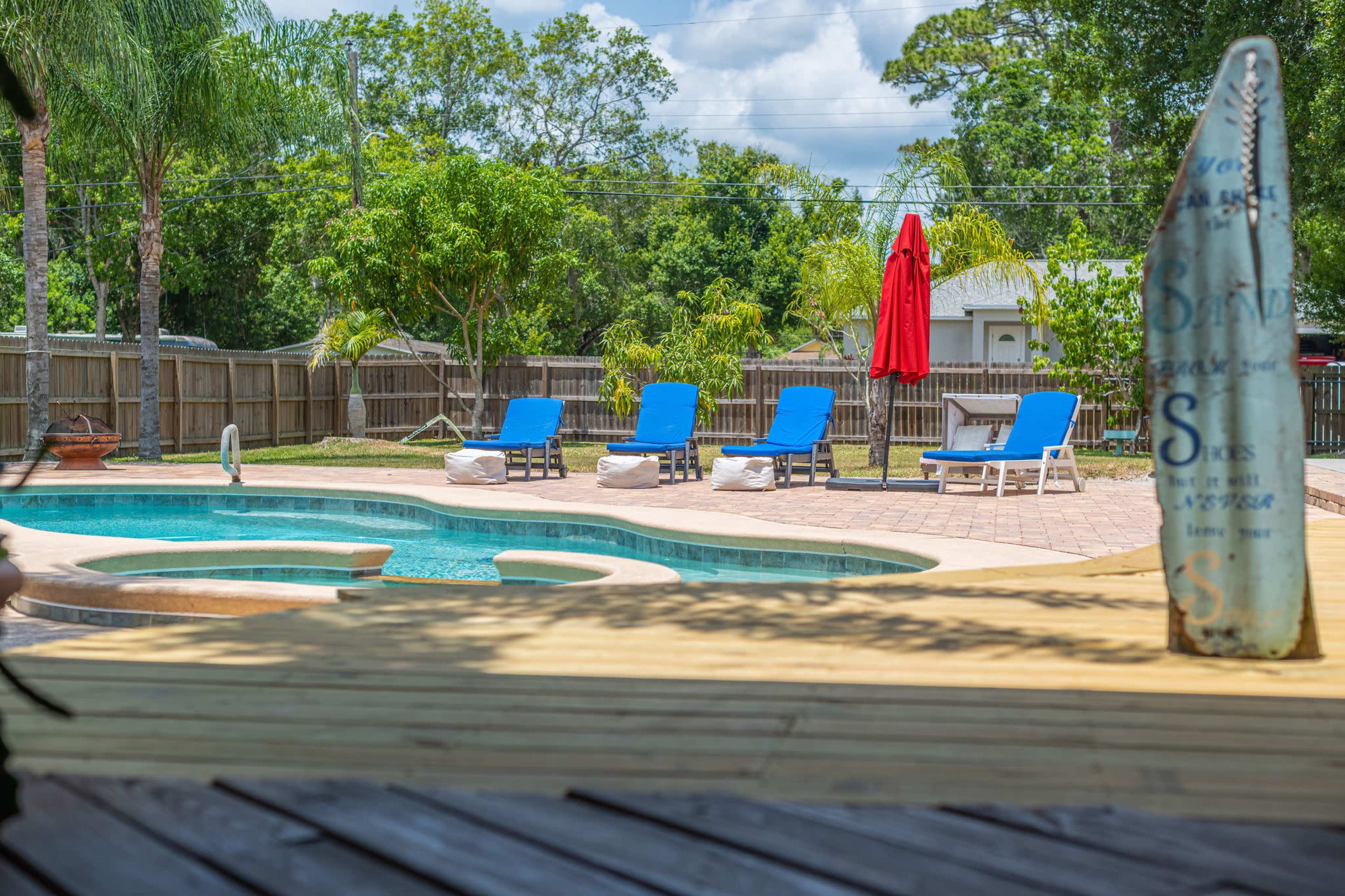 A swimming pool with lounge chairs and a red umbrella is surrounded by greenery in a fenced backyard.