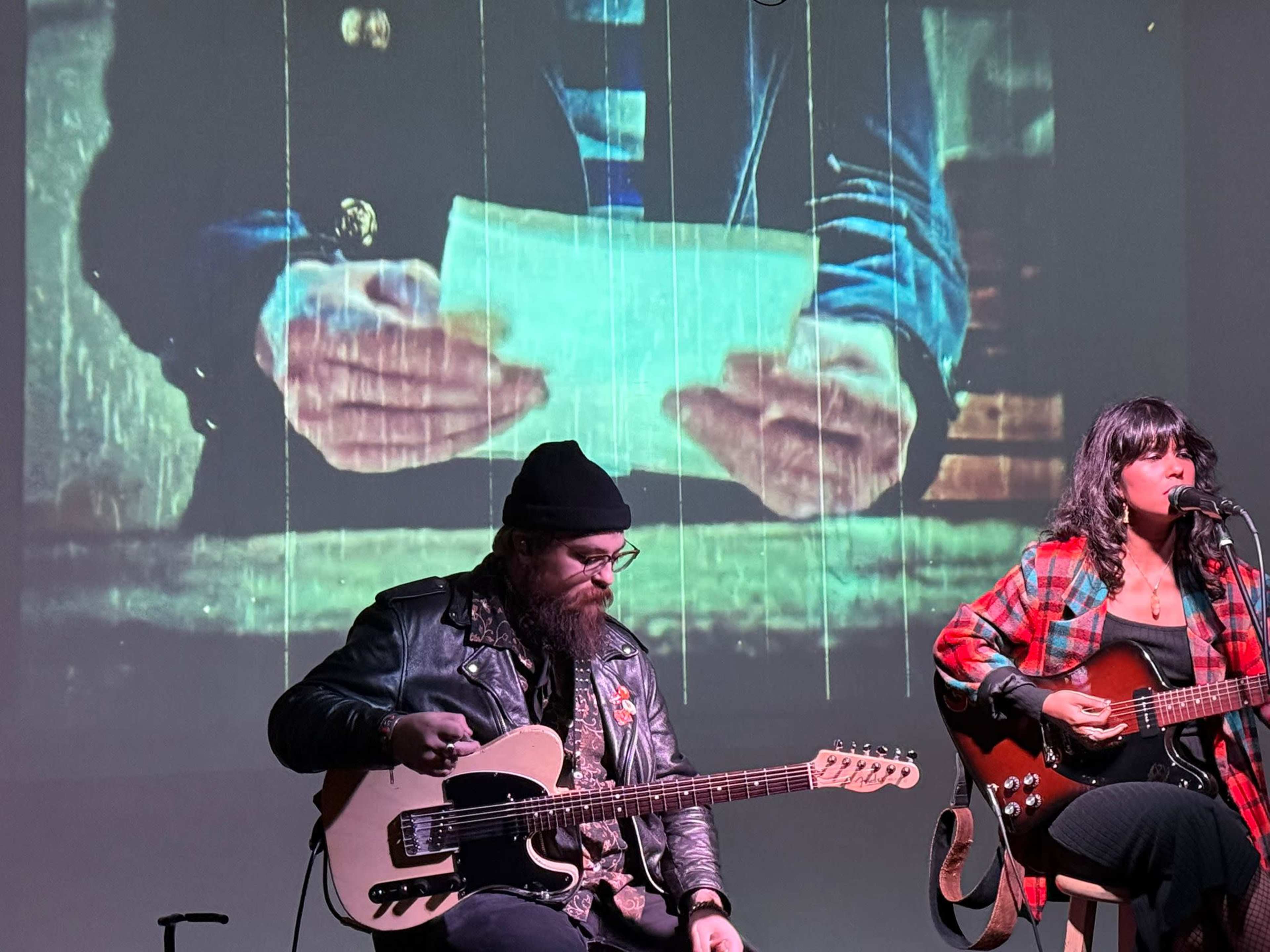 Two musicians perform with guitars in front of a projected image of hands holding a letter.