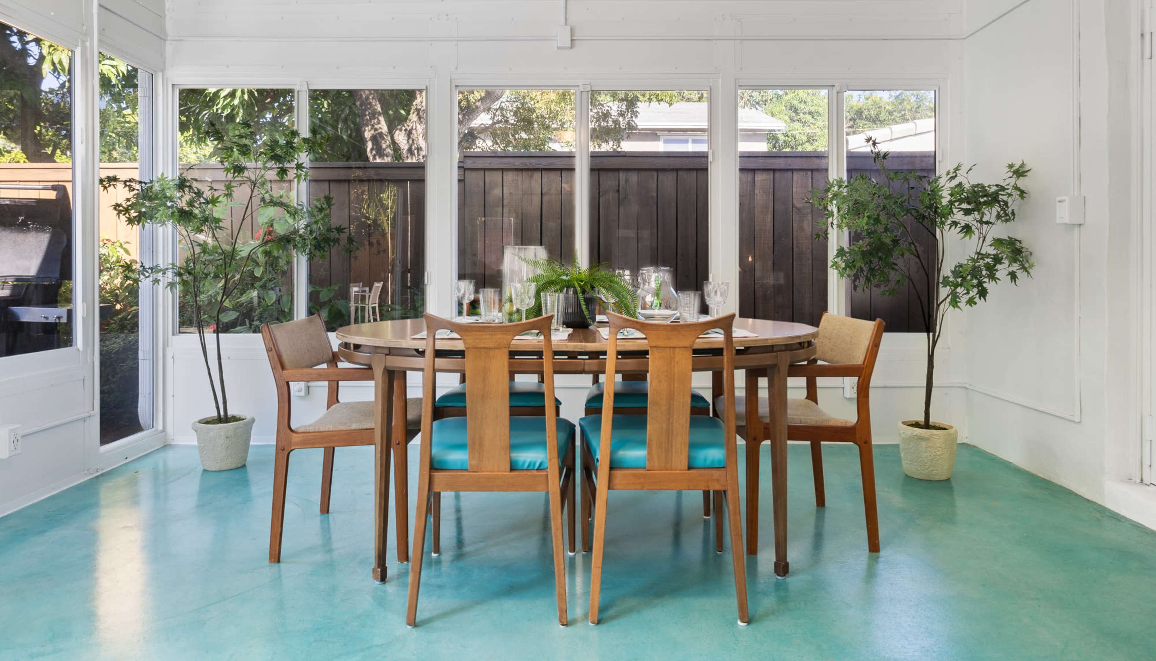 A dining table with wooden chairs and teal cushions is set in a sunlit room surrounded by large windows and potted plants.
